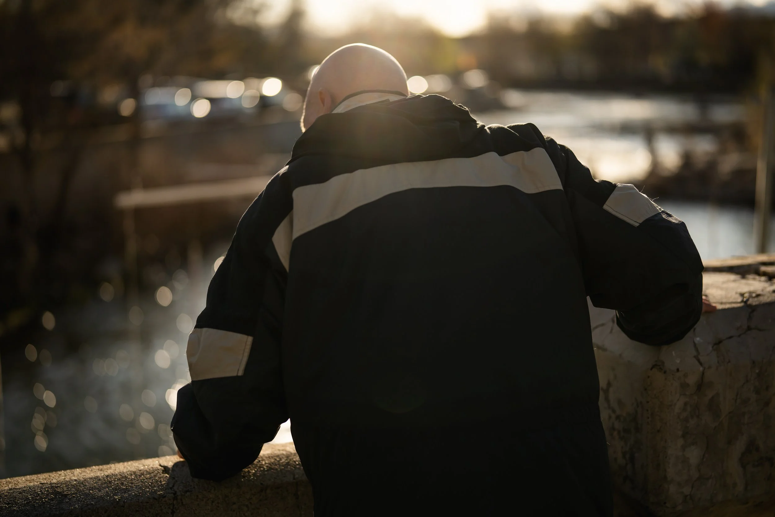 A man with a shaved head wearing a black and gray jacket leaning on a stone railing, looking down at water with the sun setting behind him.