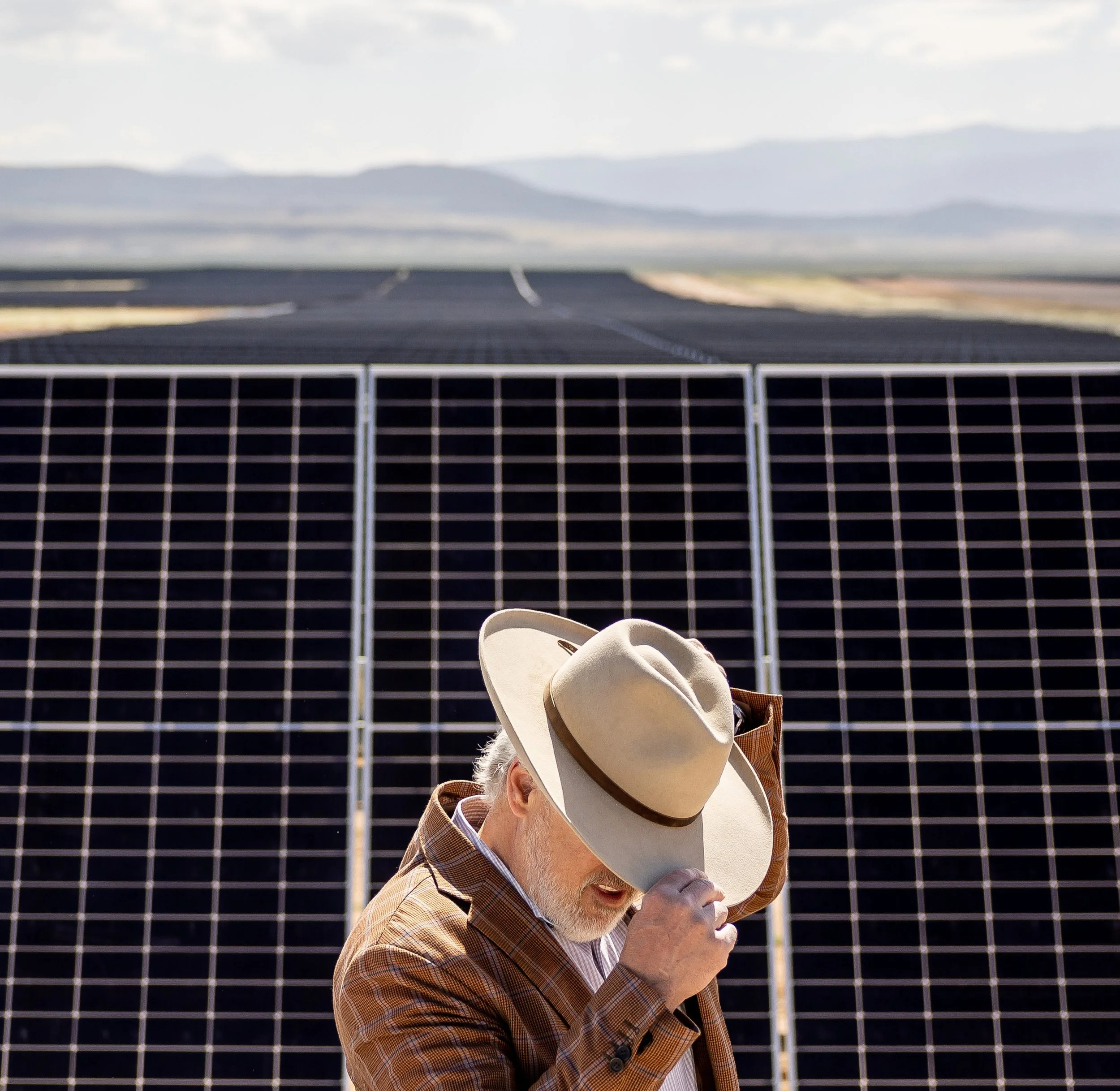 An elderly man wearing a plaid jacket and a light-colored hat adjusting his hat while standing near solar panels in a desert landscape.