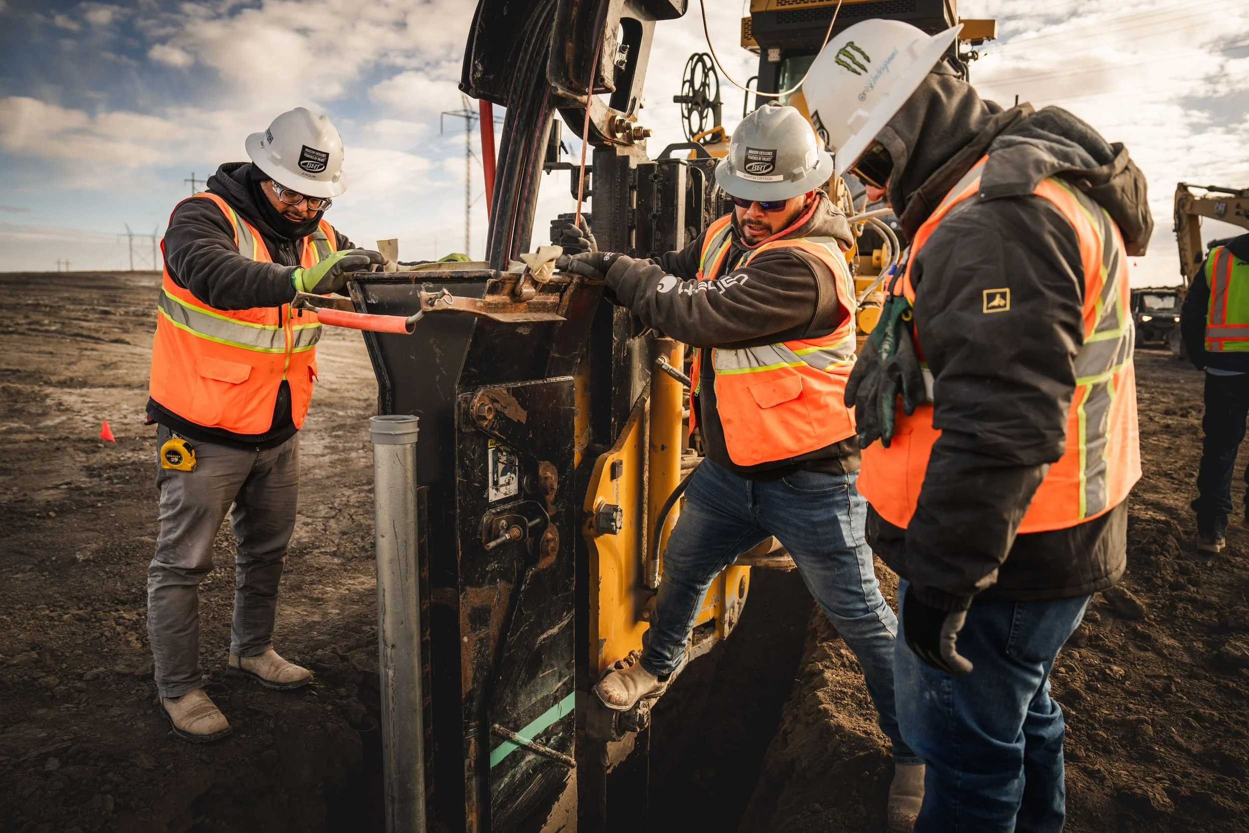 Three construction workers wearing safety vests and helmets operate heavy machinery on a dirt construction site during daytime.