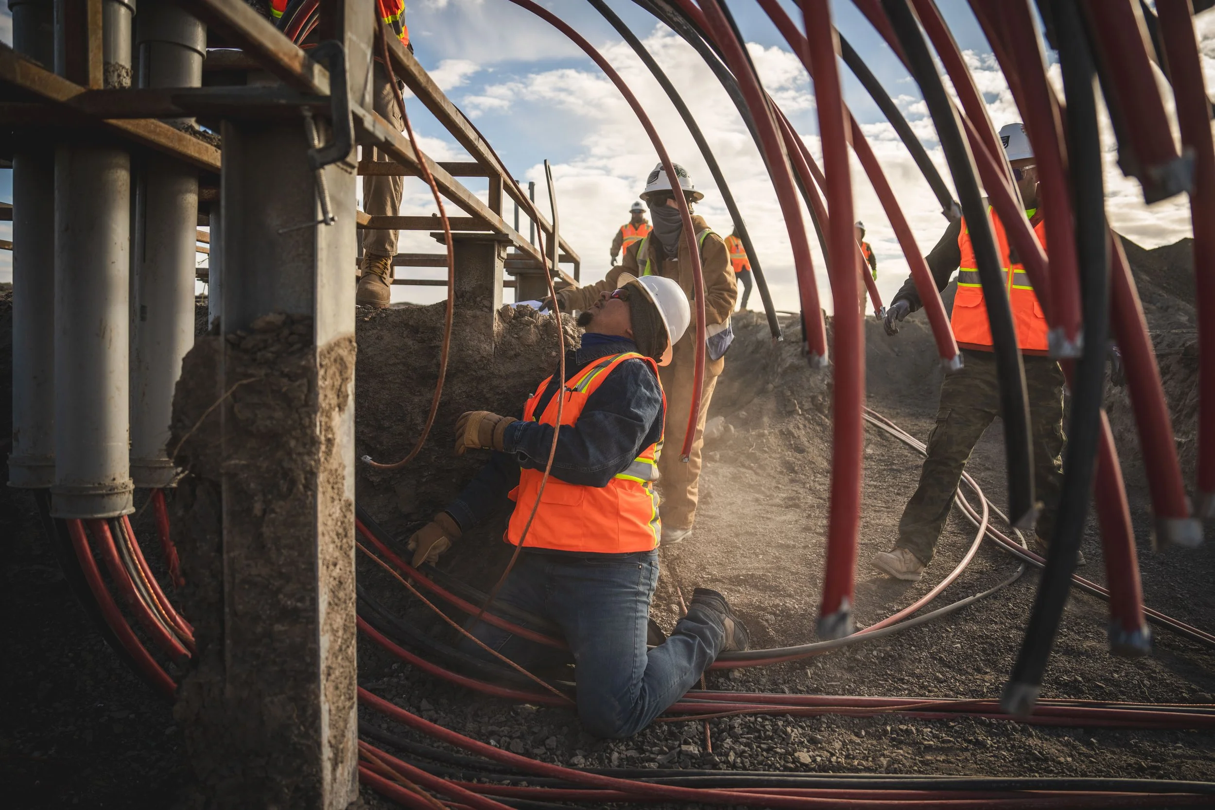 Construction workers wearing safety gear, working under a bridge or structure with exposed cables and support beams during daytime.