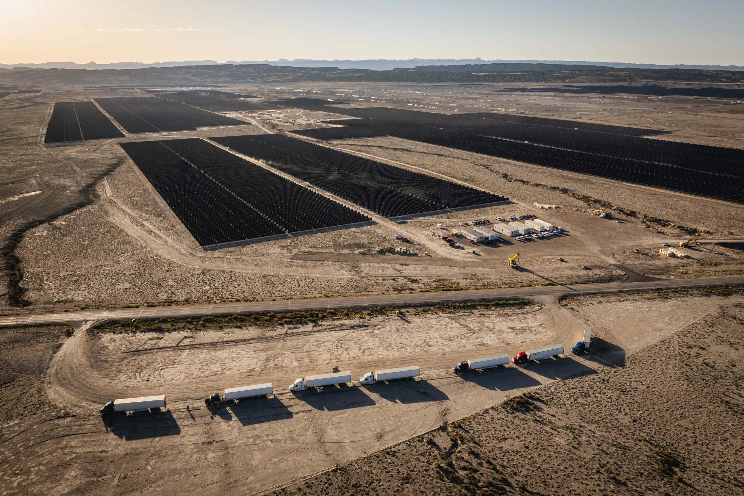 Aerial view of a solar farm with rows of large solar panels in a desert landscape, a truck convoy on a dirt road, and support buildings nearby under a clear sky.