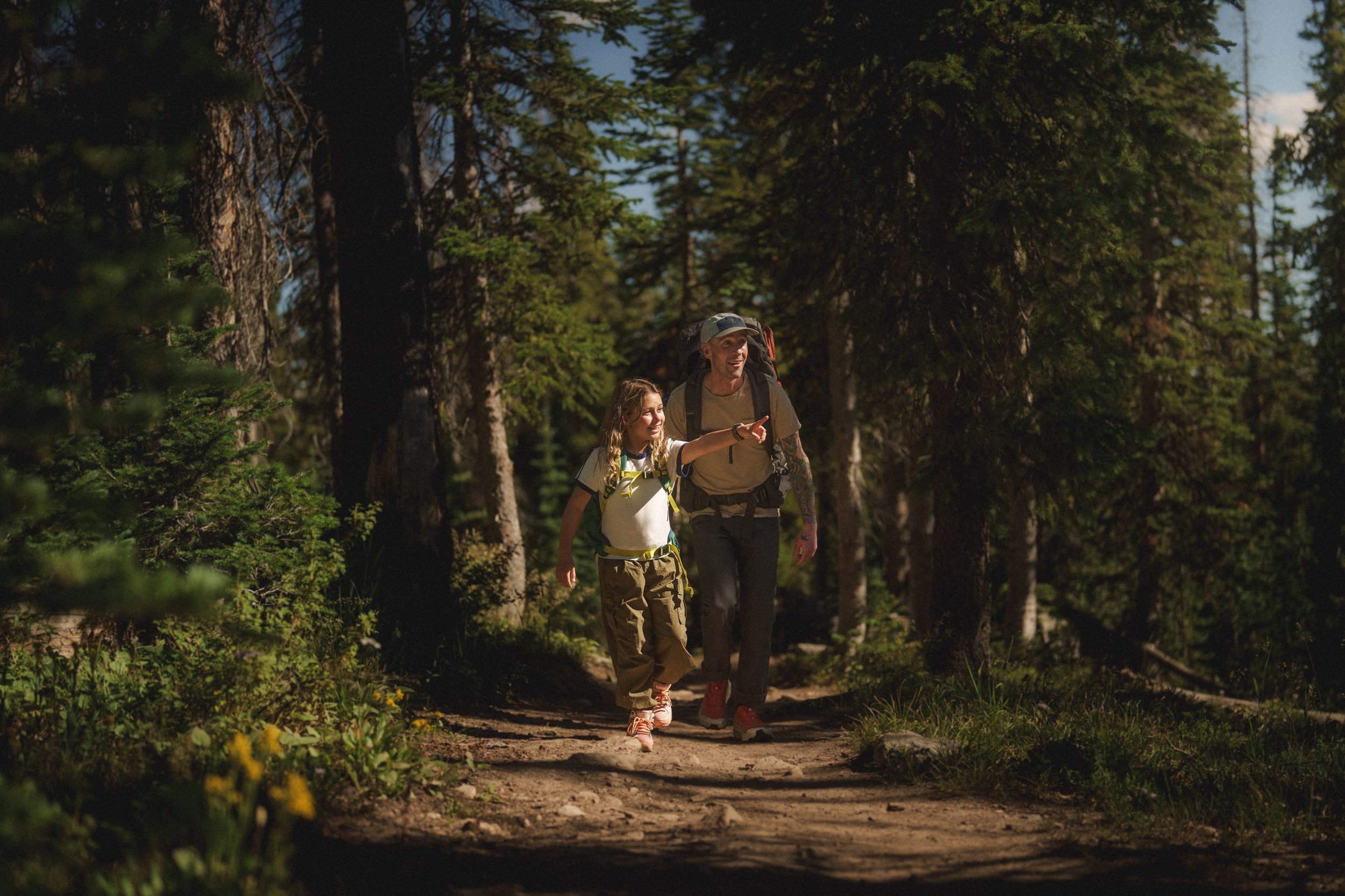 A man and a girl hiking on a trail in a forest, with tall trees and sunlight filtering through the leaves.