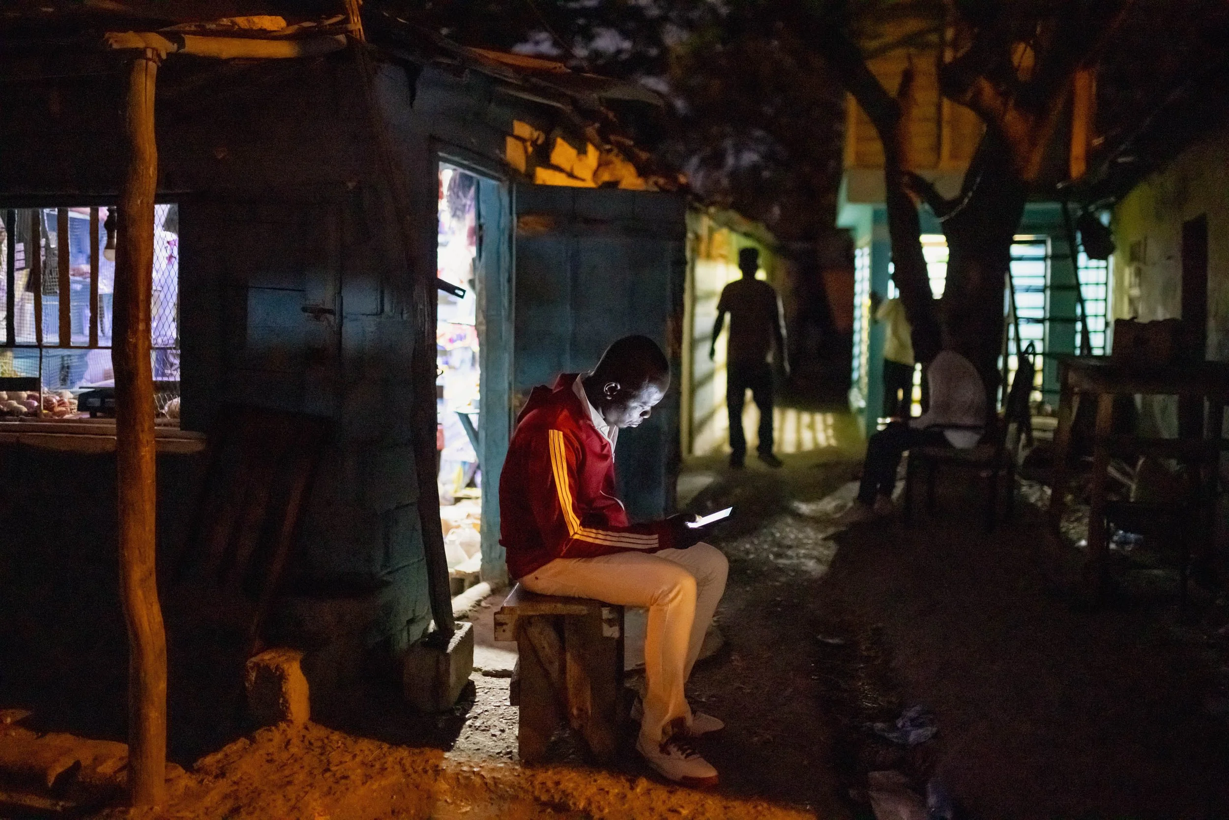 A young man in a red jacket and beige pants sitting on a small wooden bench outside a shop at night, illuminated by the light from his phone. In the background, there are other people, trees, and rustic houses, with a dirt path and furniture.