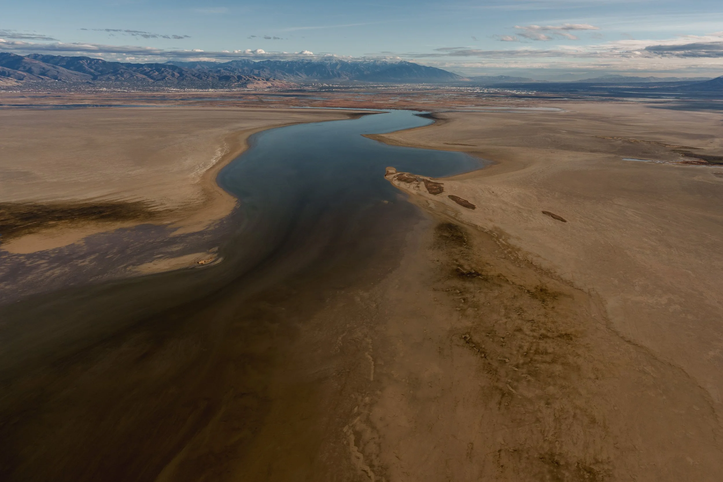 A winding river flowing through a dry, arid landscape with mountains in the distance and a partly cloudy sky.