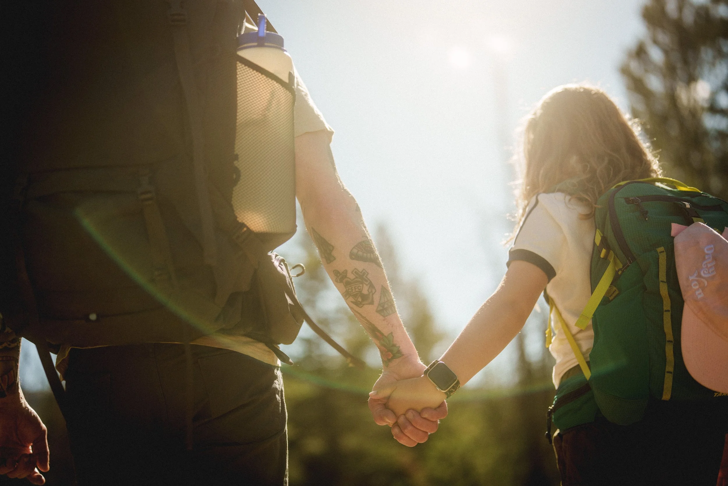 A person and a child holding hands outdoors in sunlight, both wearing backpacks.