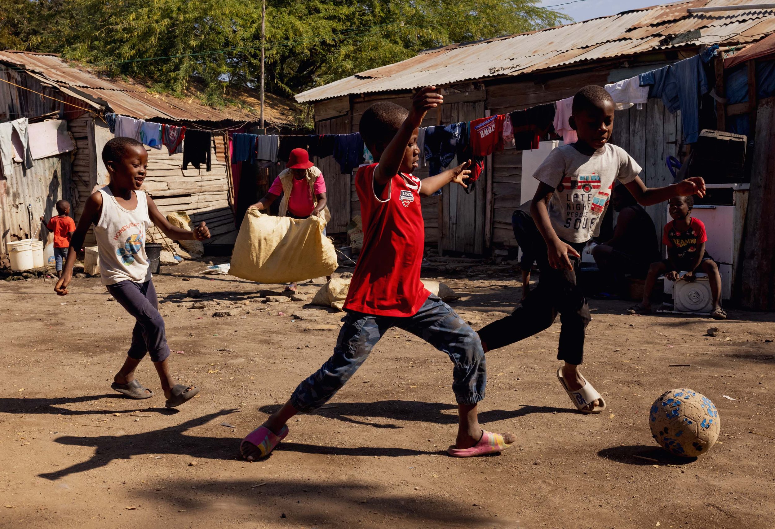 Children playing soccer on a dirt ground near wooden houses with laundry hanging on clotheslines.