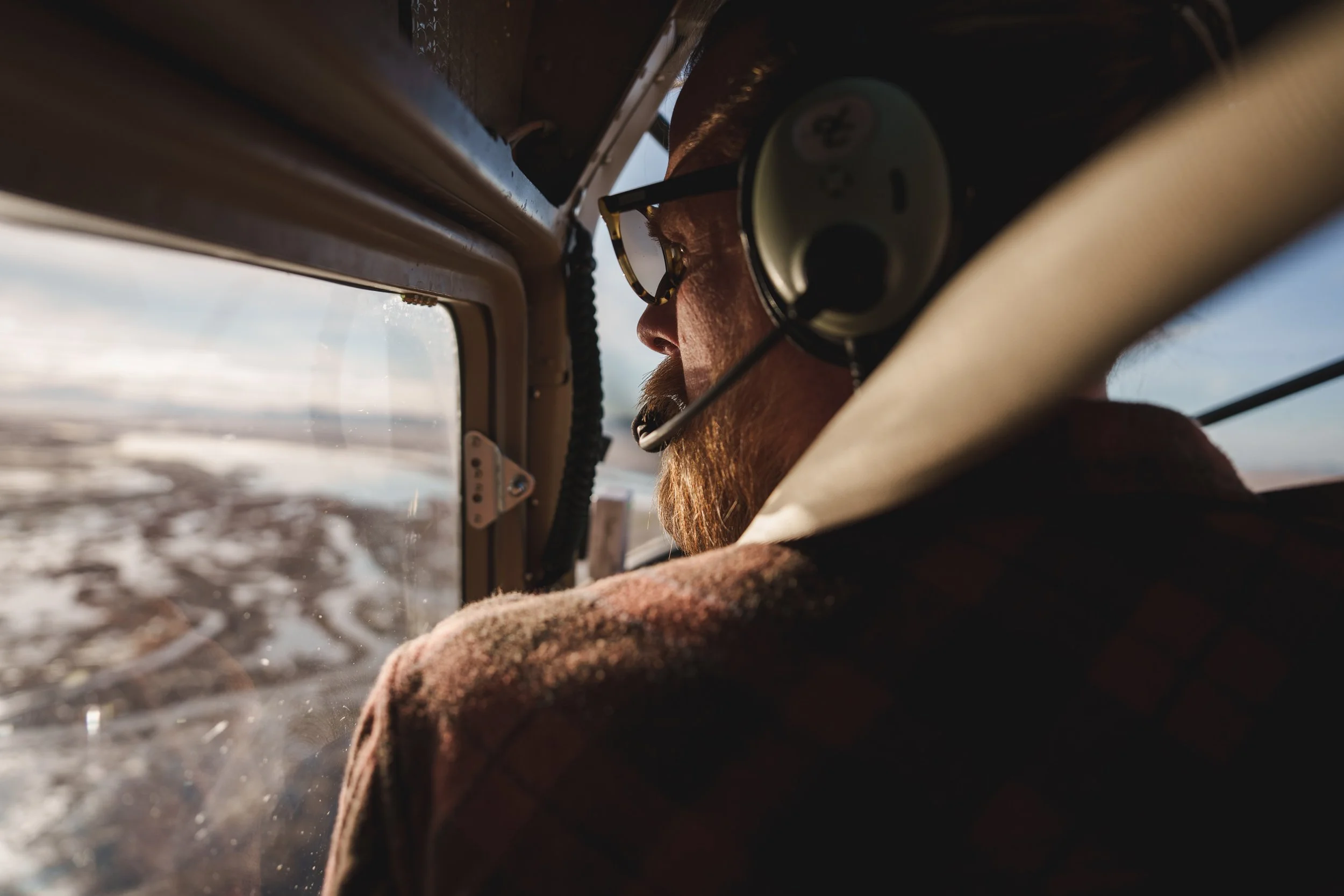 A man flying a small aircraft, wearing headphones and glasses, looking out of the cockpit at the landscape below.