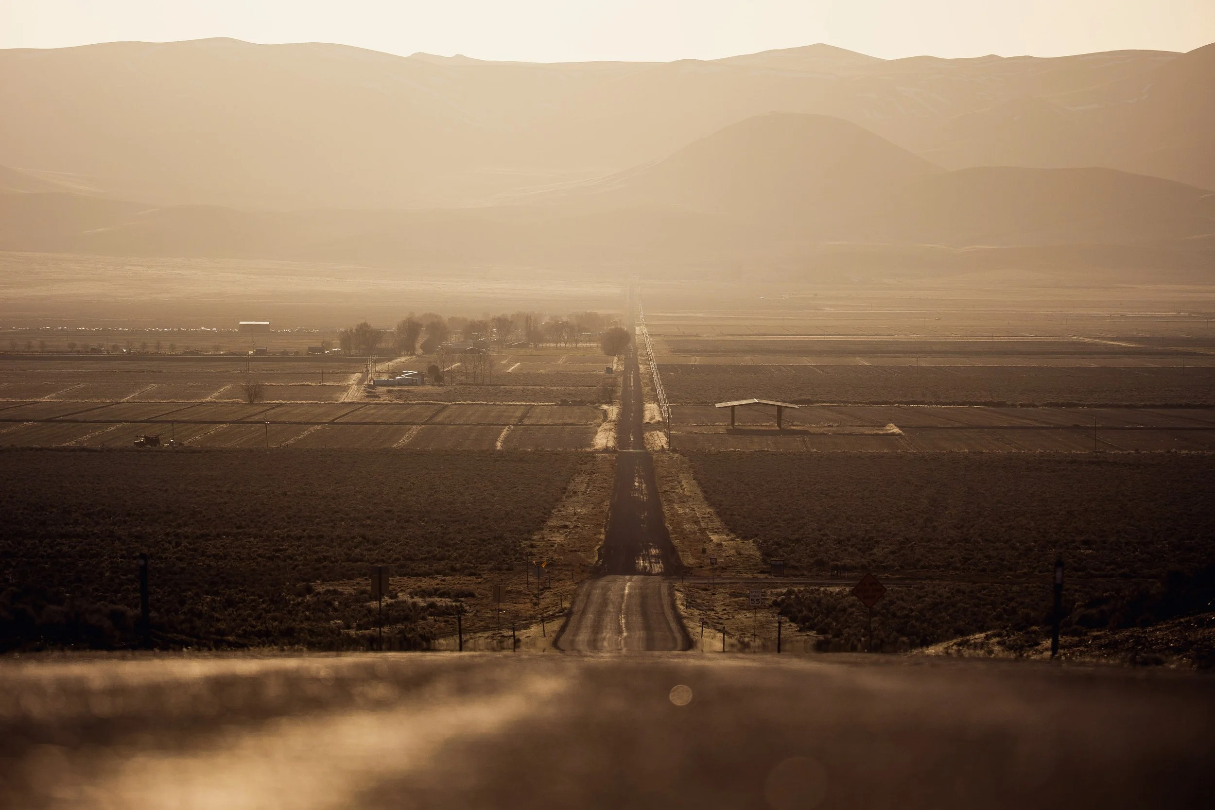 Long straight dirt road stretching into the distance through a rural landscape with fields and a few scattered buildings, with mountains in the background and a hazy sky.