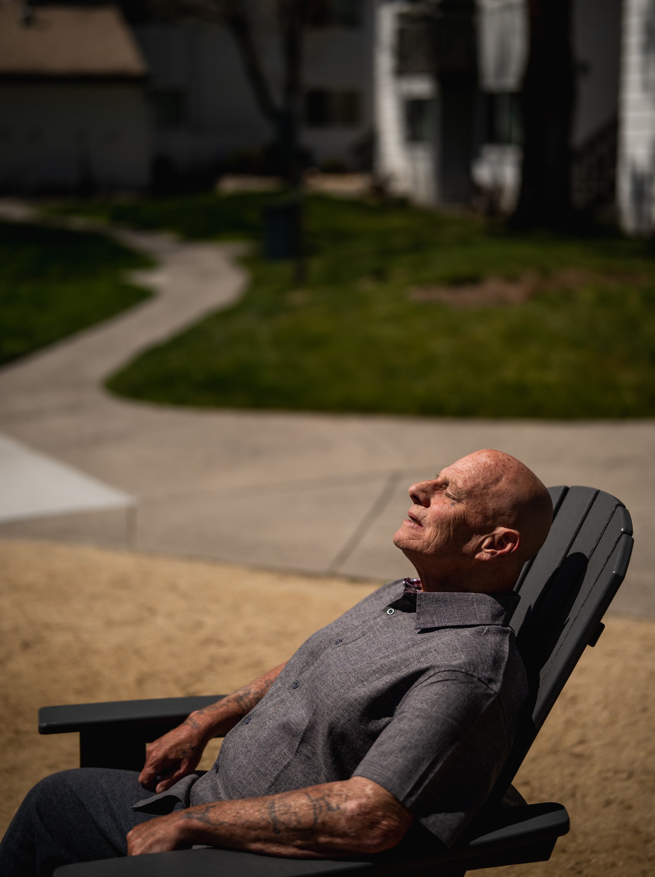 An elderly man with tattoos on his arms, wearing a gray shirt, is relaxing in a black outdoor chair with his eyes closed and face tilted upward, soaking up the sun.