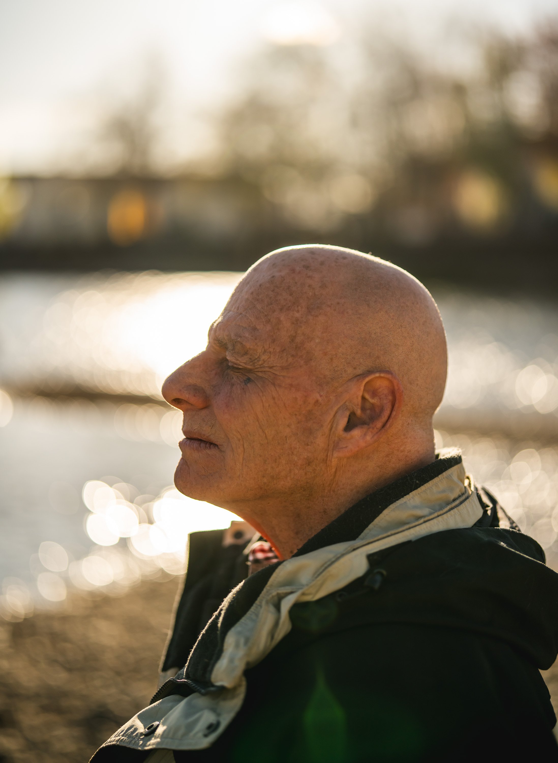 A bald man with wrinkled skin sitting outdoors near water, eyes closed, in profile view, with sunlight reflecting on the water in the background.