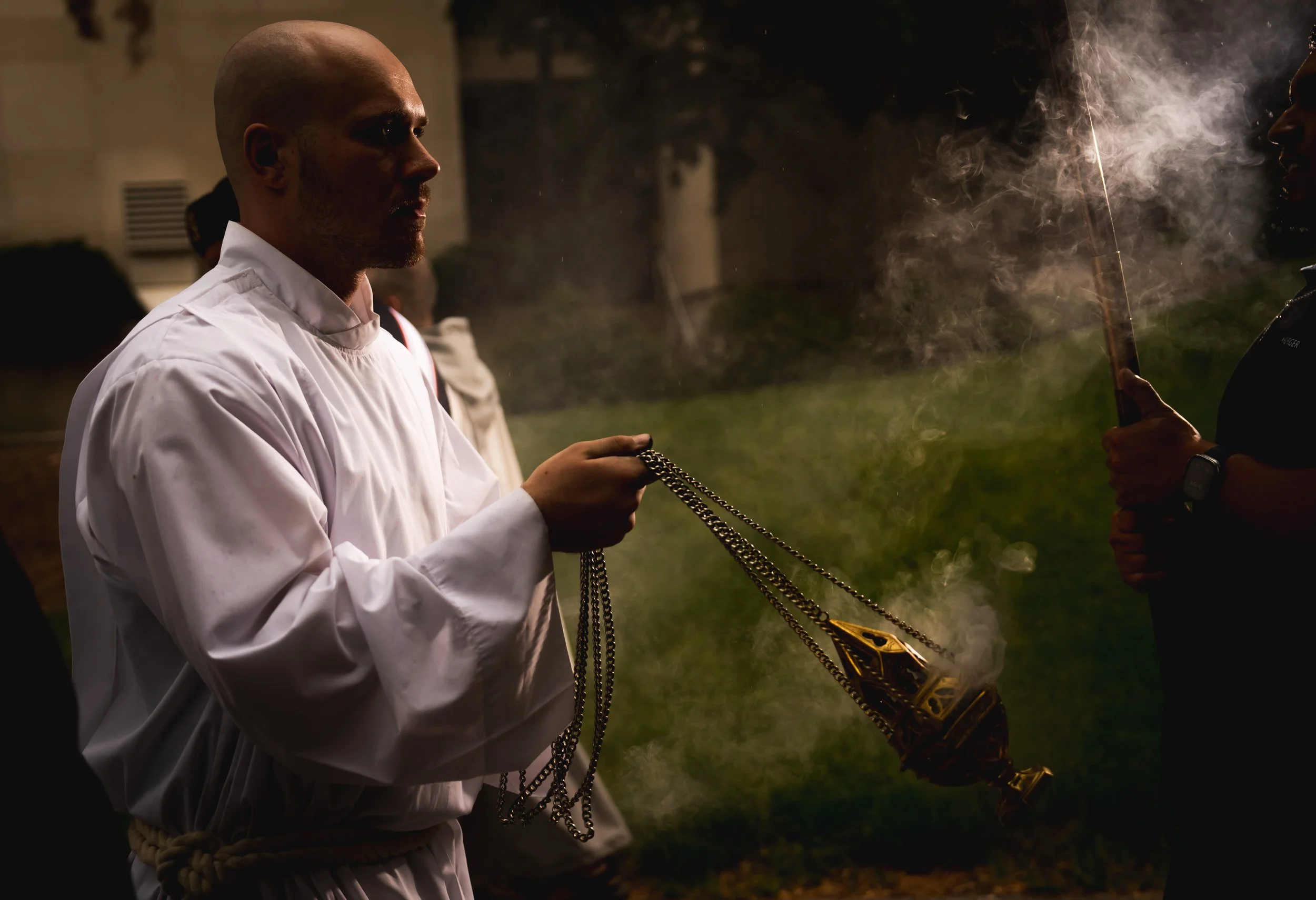 A man dressed in white holding chains attached to a burning censer, standing outdoors at night with smoke in the air.