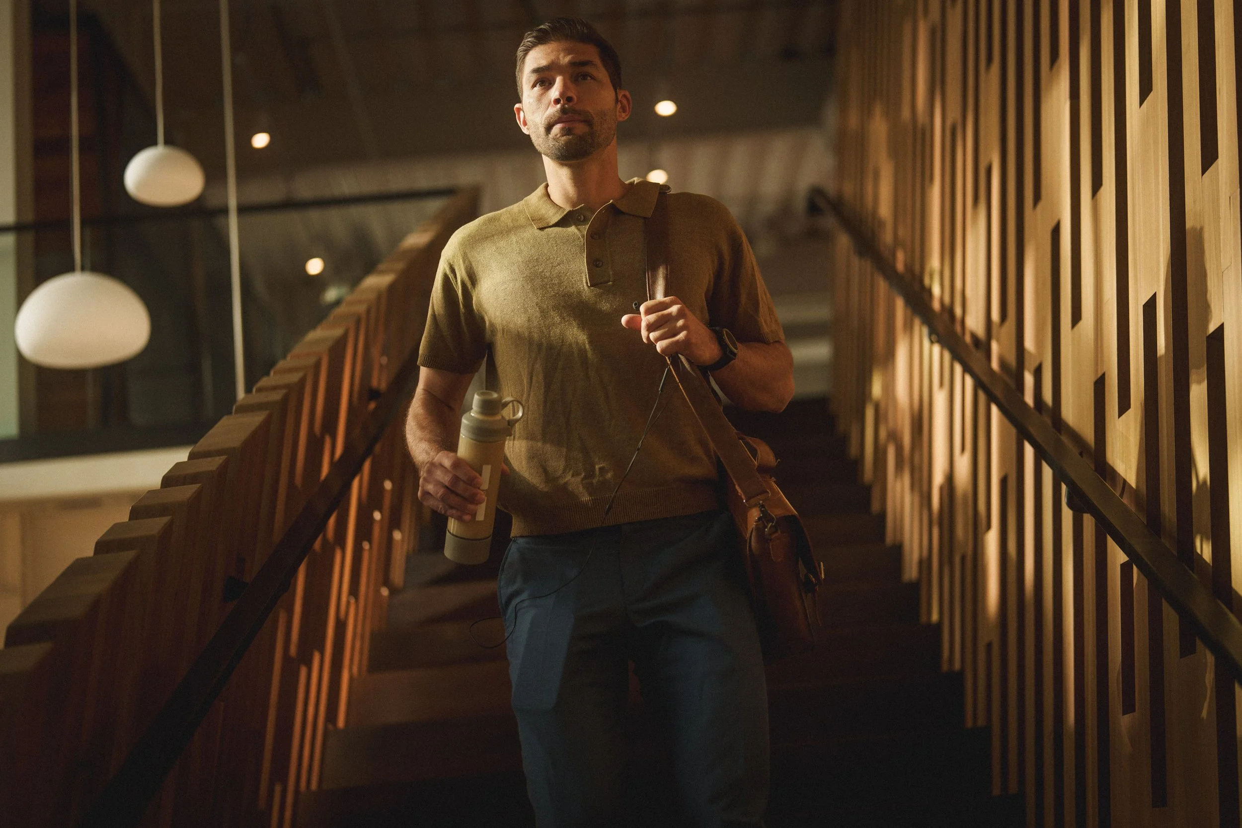 A man with a beard, wearing a brown polo shirt, walking up a staircase inside a modern building, holding a water bottle and carrying a shoulder bag.