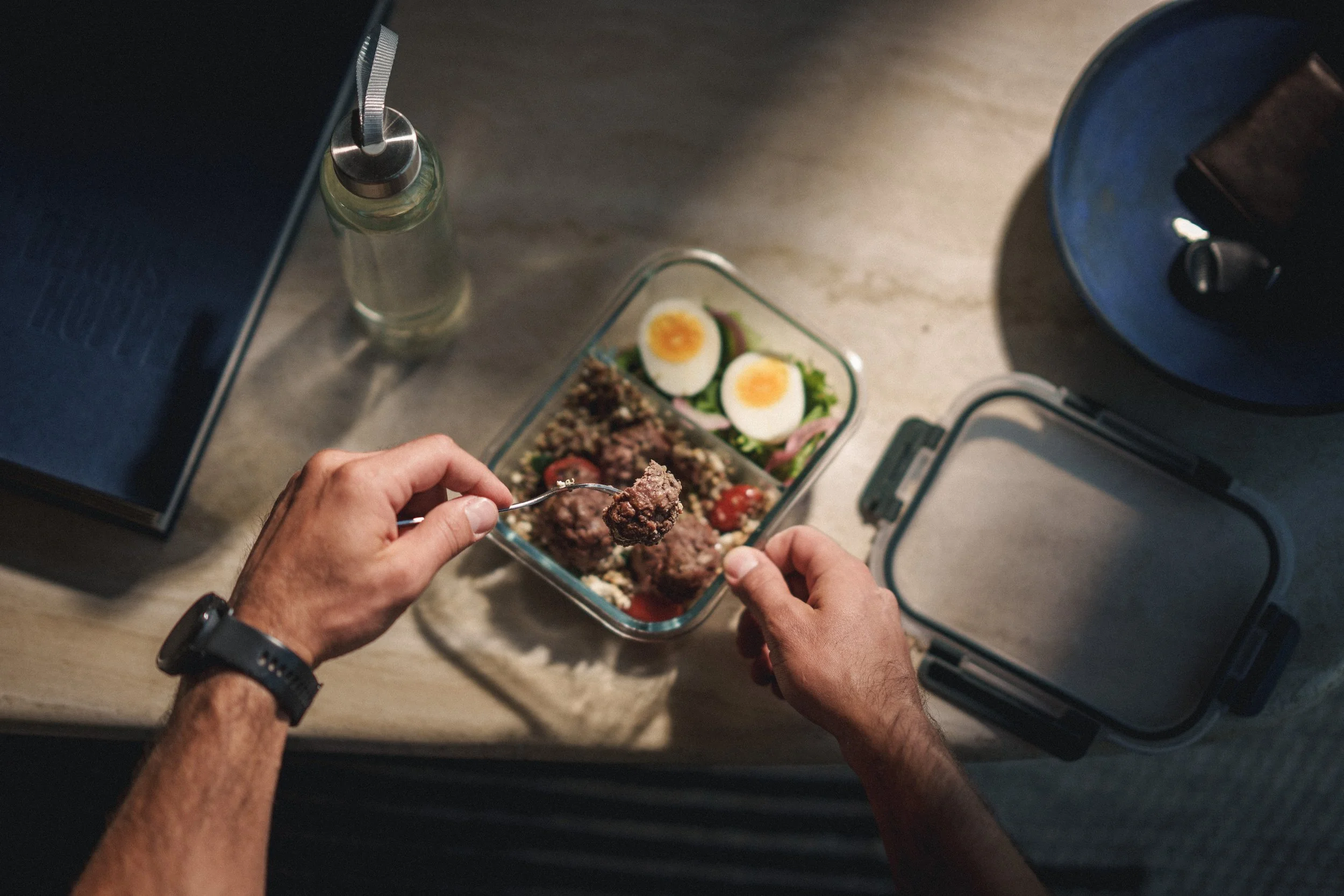 Person holding a fork with a meatball, lunch container with salad and quail eggs, a water bottle, and a rice cooker on a table.
