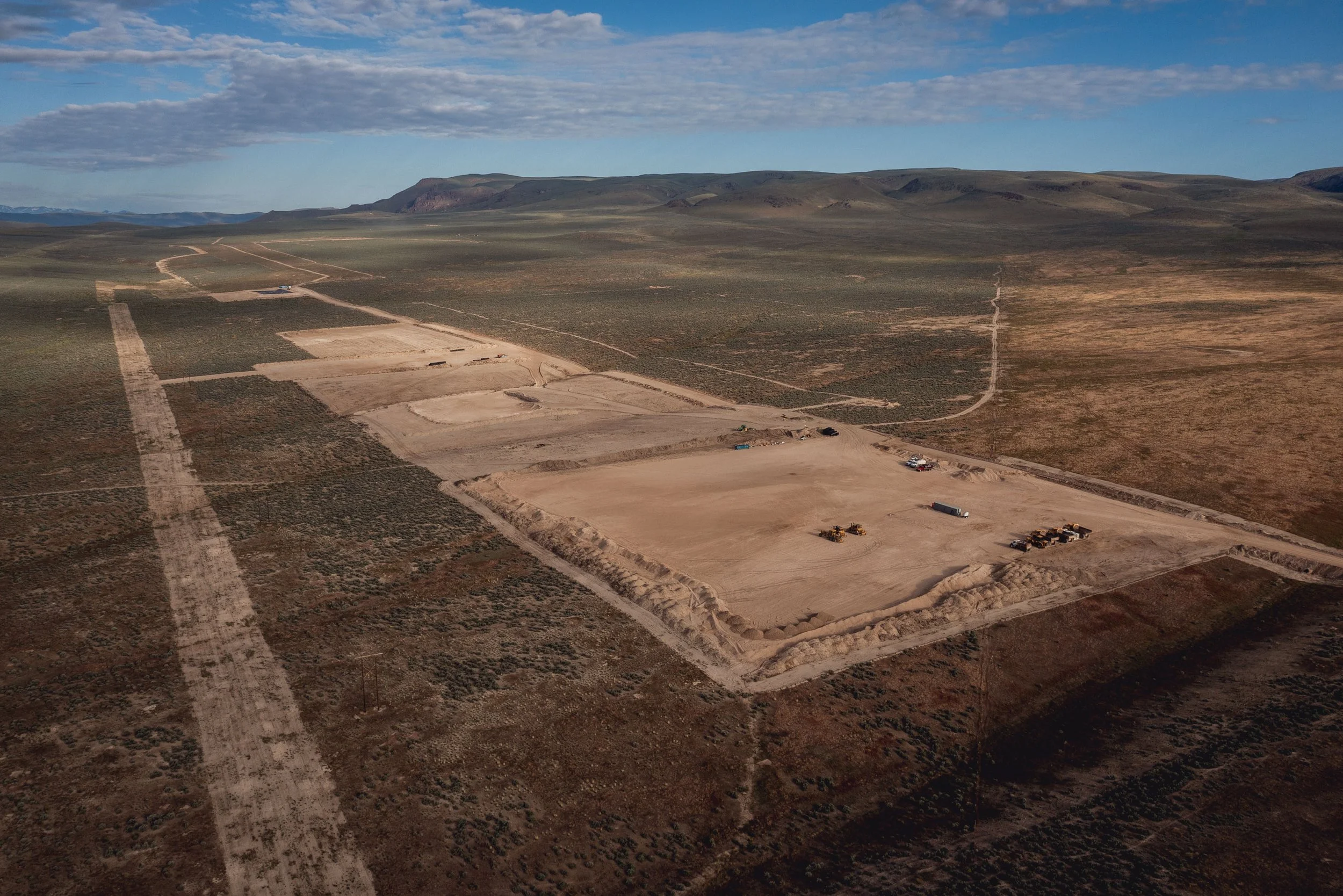 An aerial view of a large construction site in a desert landscape, with multiple cleared areas, construction vehicles, and equipment, surrounded by open land and distant mountains under a partly cloudy sky.