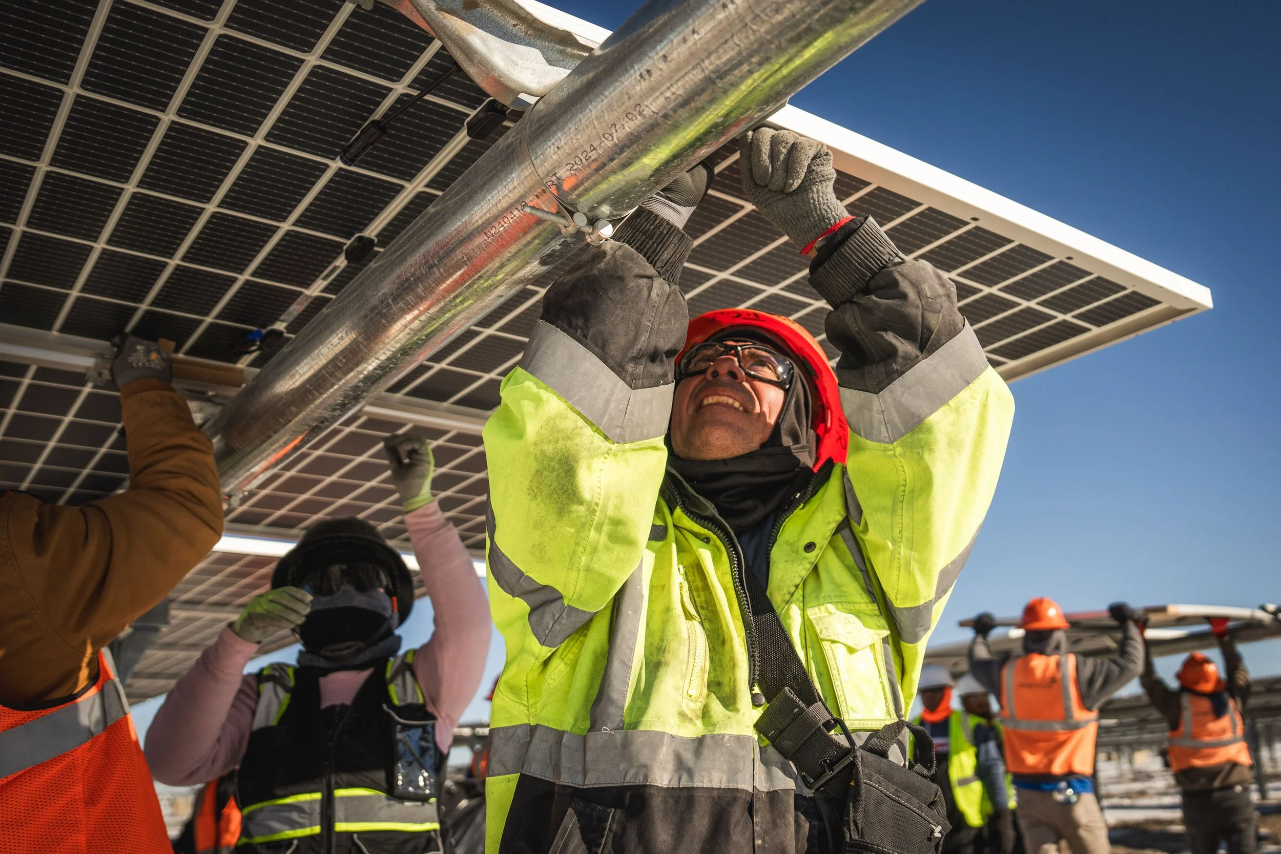 Workers installing solar panels on a rooftop during daytime, wearing safety gear including helmets and reflective vests.