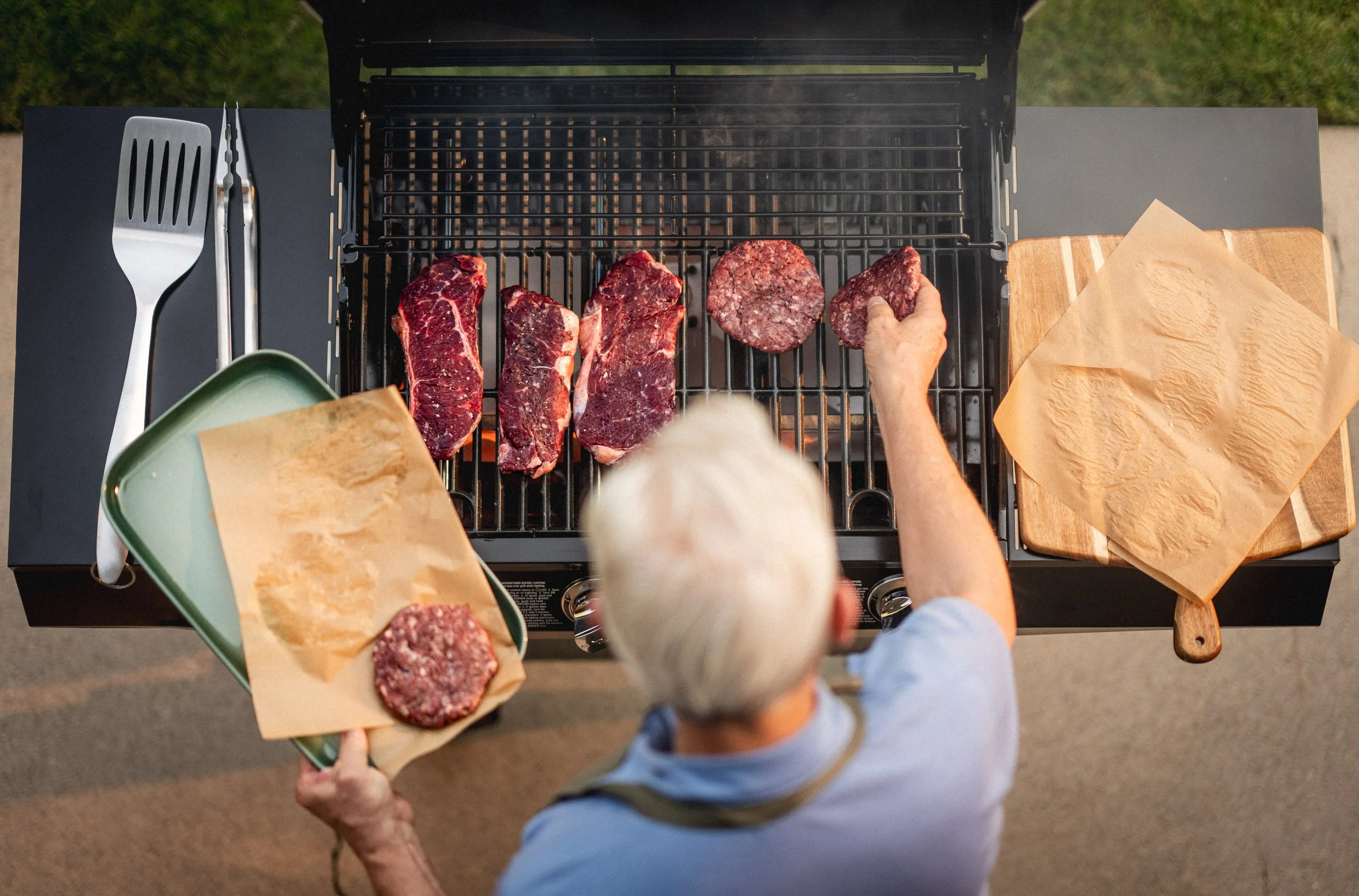 An overhead view of a man grilling various cuts of beef on a barbecue, with platters of raw meat ready to cook nearby.