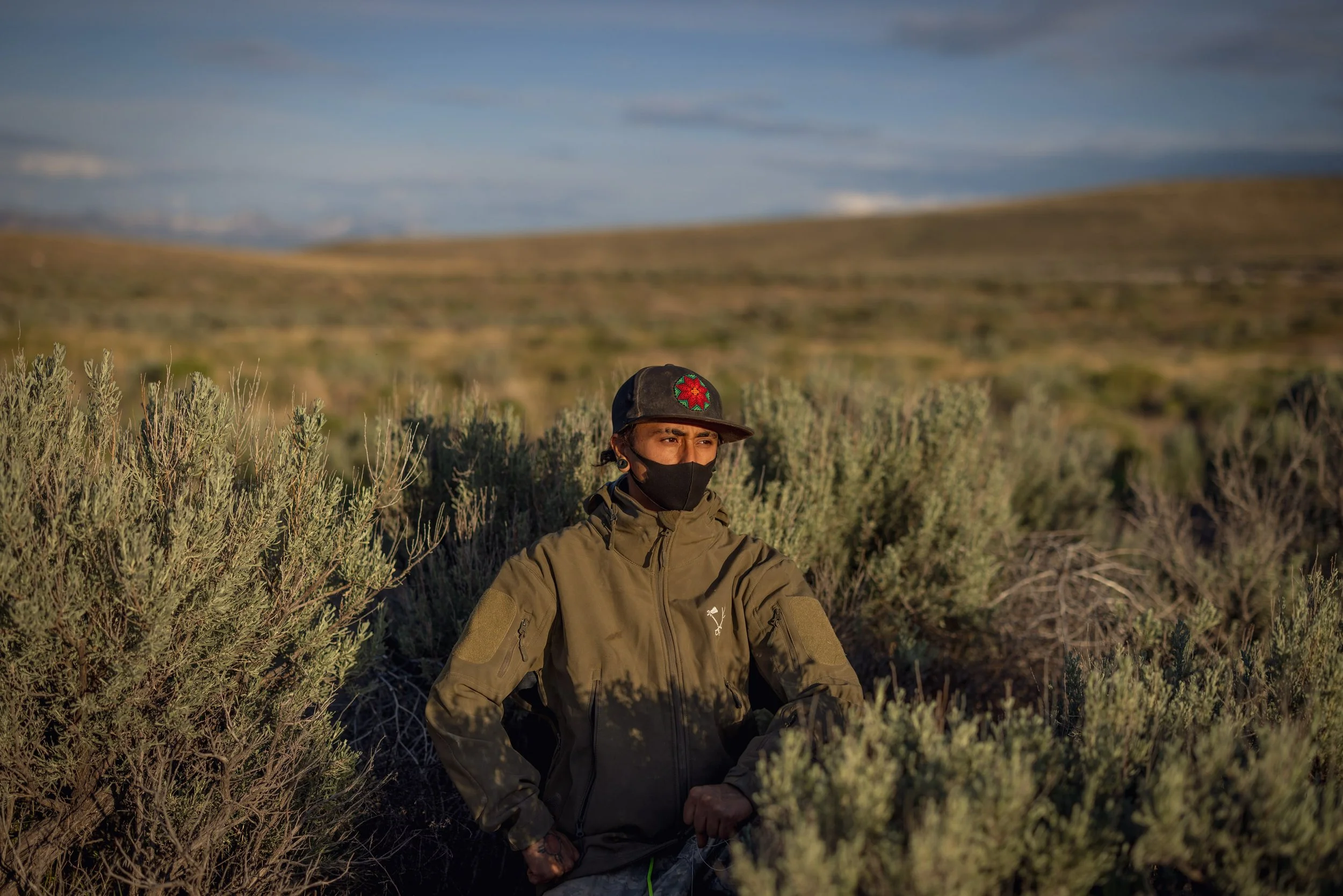 A person wearing a black face mask, a green jacket, and a black cap with a red star emblem, standing in a dry, grassy landscape with bushes.