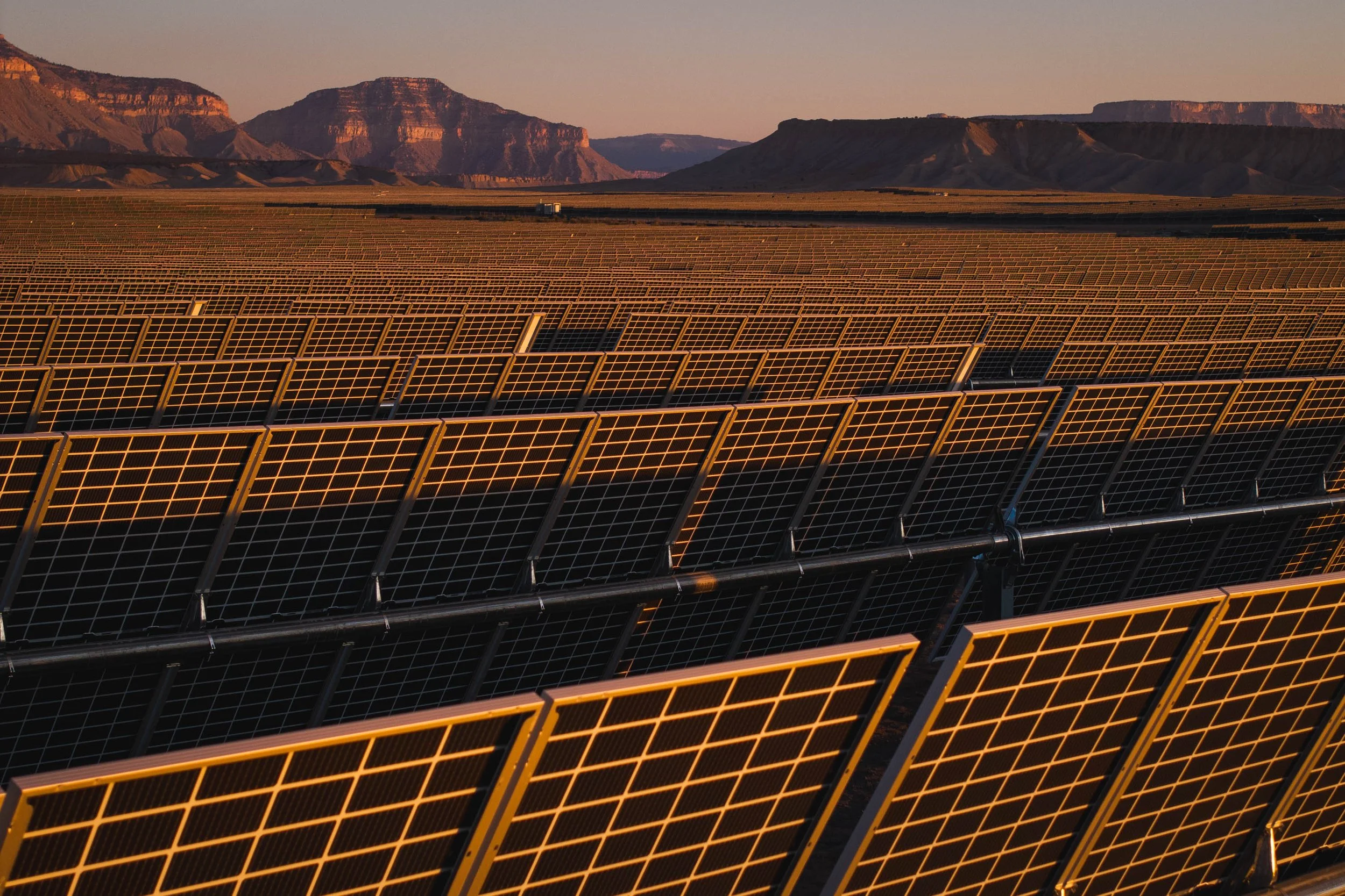 A solar farm with multiple rows of solar panels in a desert landscape, mountains in the background at sunset.