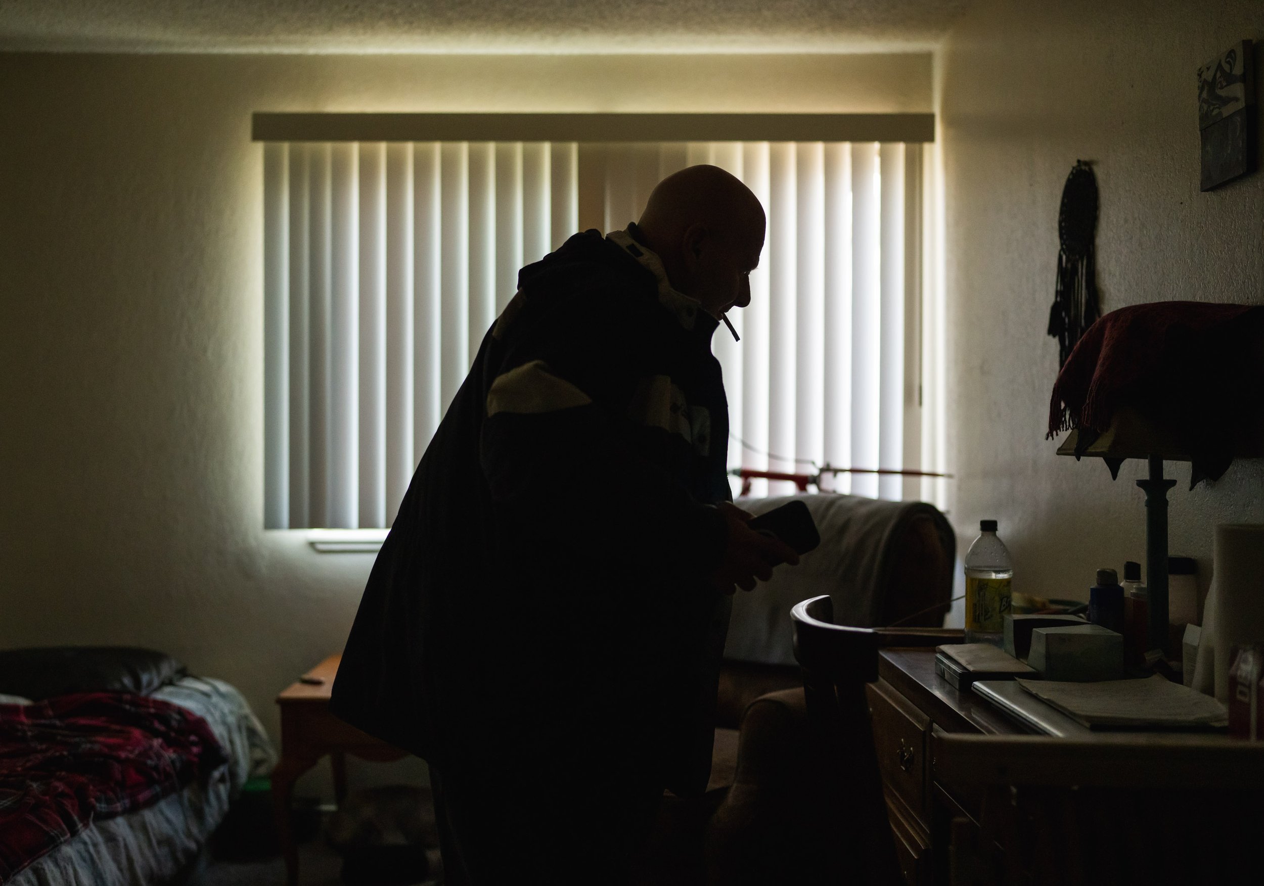Silhouette of a person with a shaved head standing in a dimly lit room, looking down at a cellphone in front of a window with closed vertical blinds.