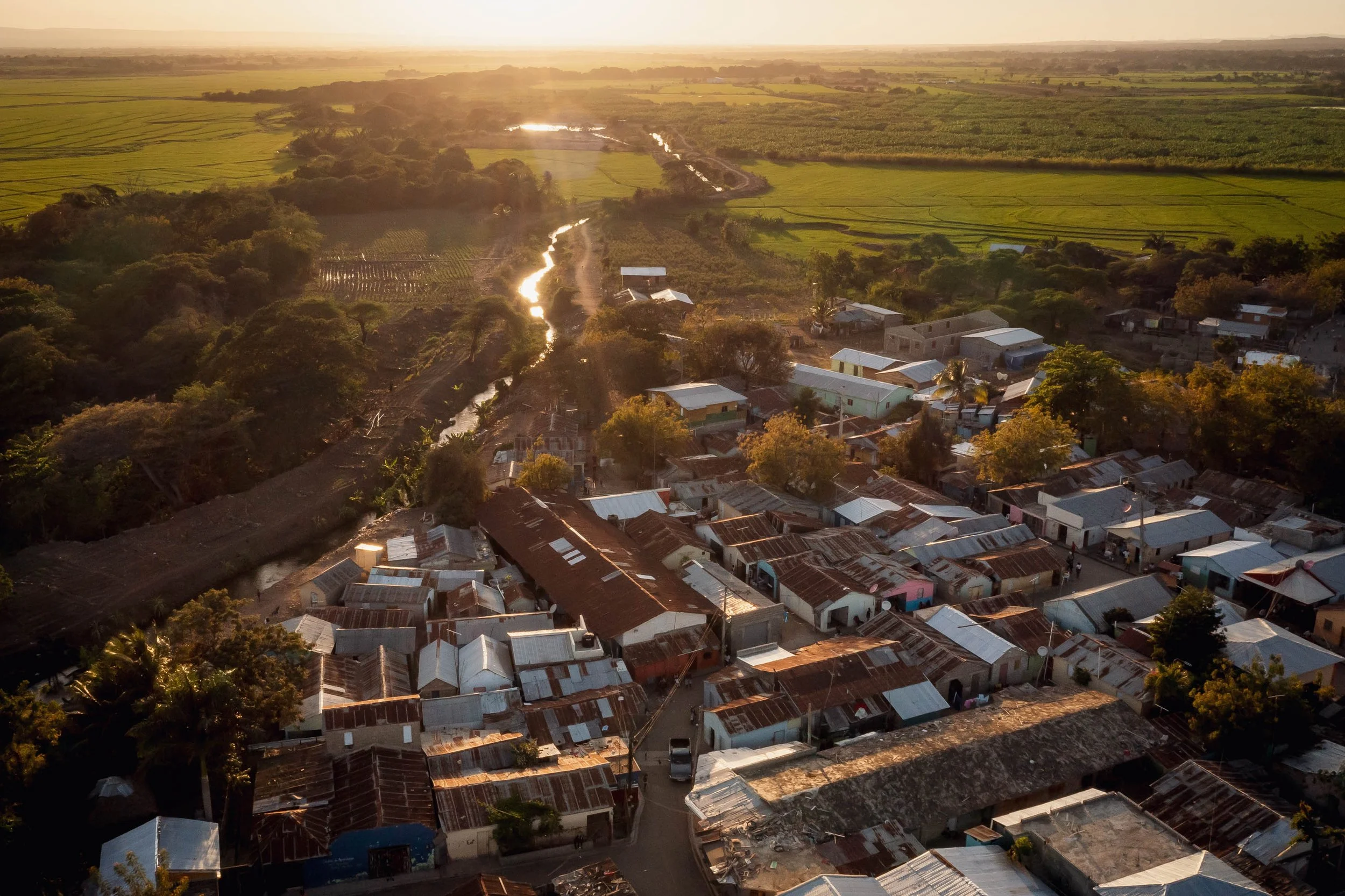 Aerial view of a small town with metal roofs, surrounded by lush green fields and trees, during sunset.
