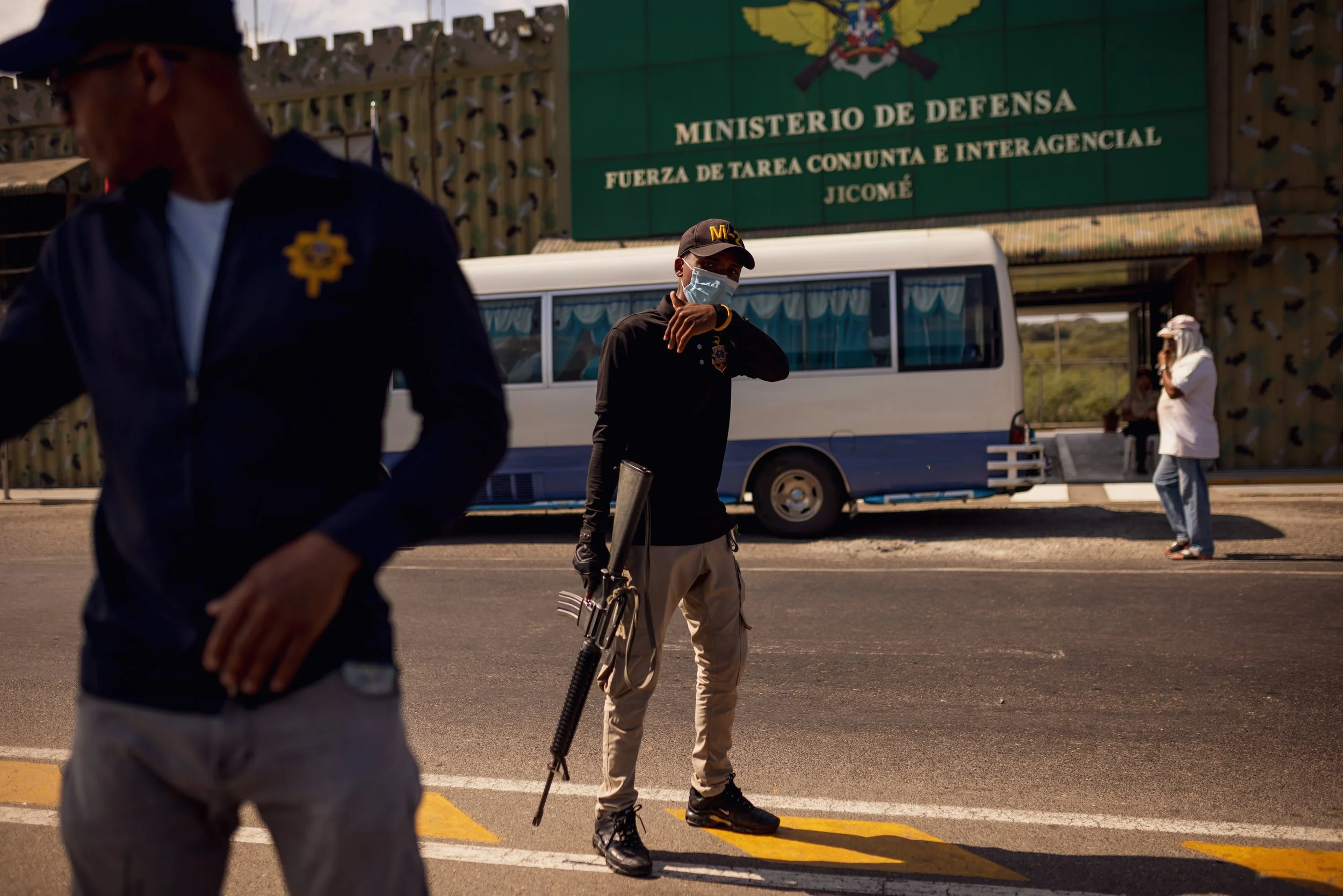 Military or security personnel standing on the street, with a bus and a building behind them that has a sign reading 'MINISTERIO DE DEFENSA' and other Spanish text. The personnel are wearing face masks, and one is holding a firearm.