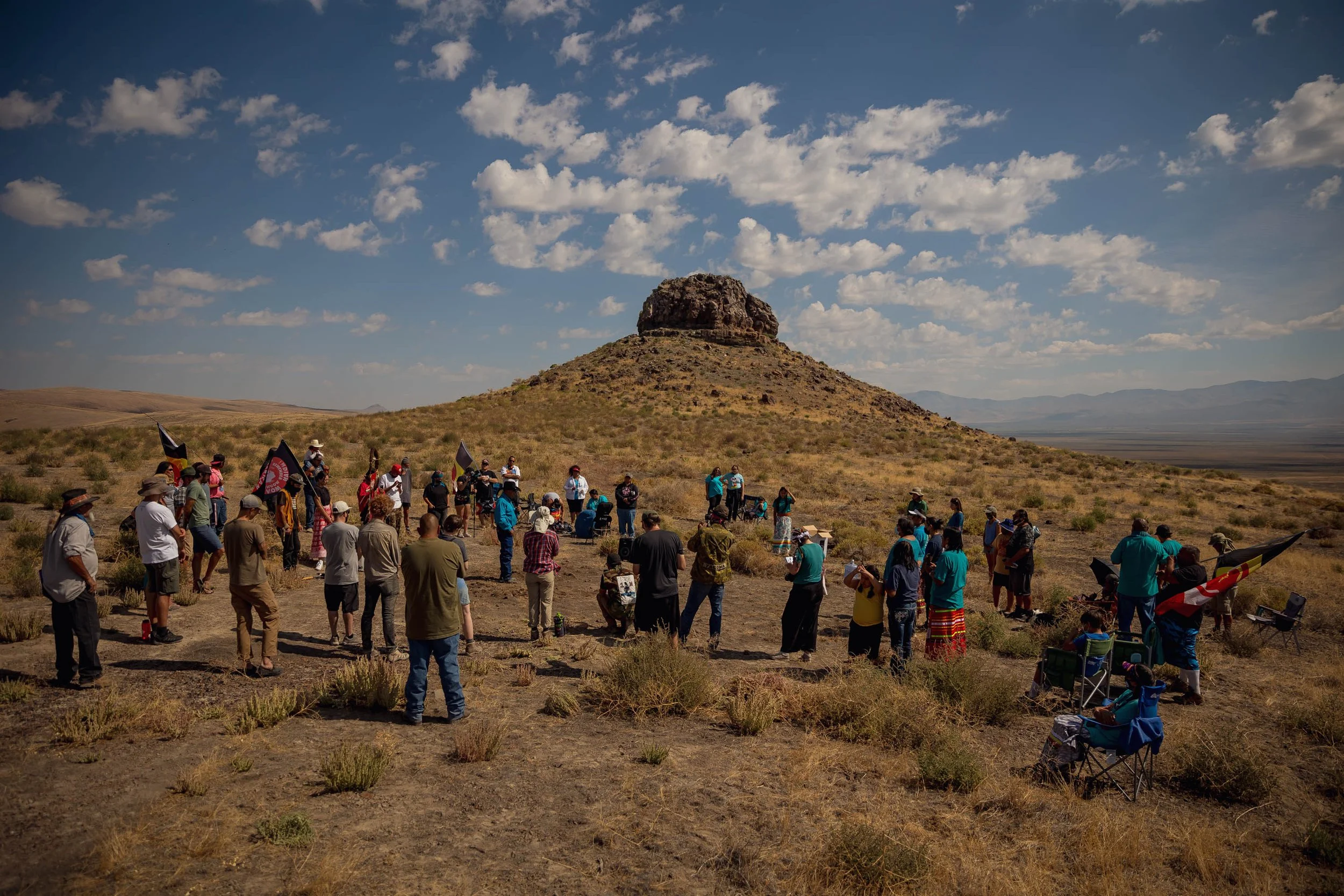 Group of people gathered outdoors in a desert landscape, facing a large rock formation under a partly cloudy sky.