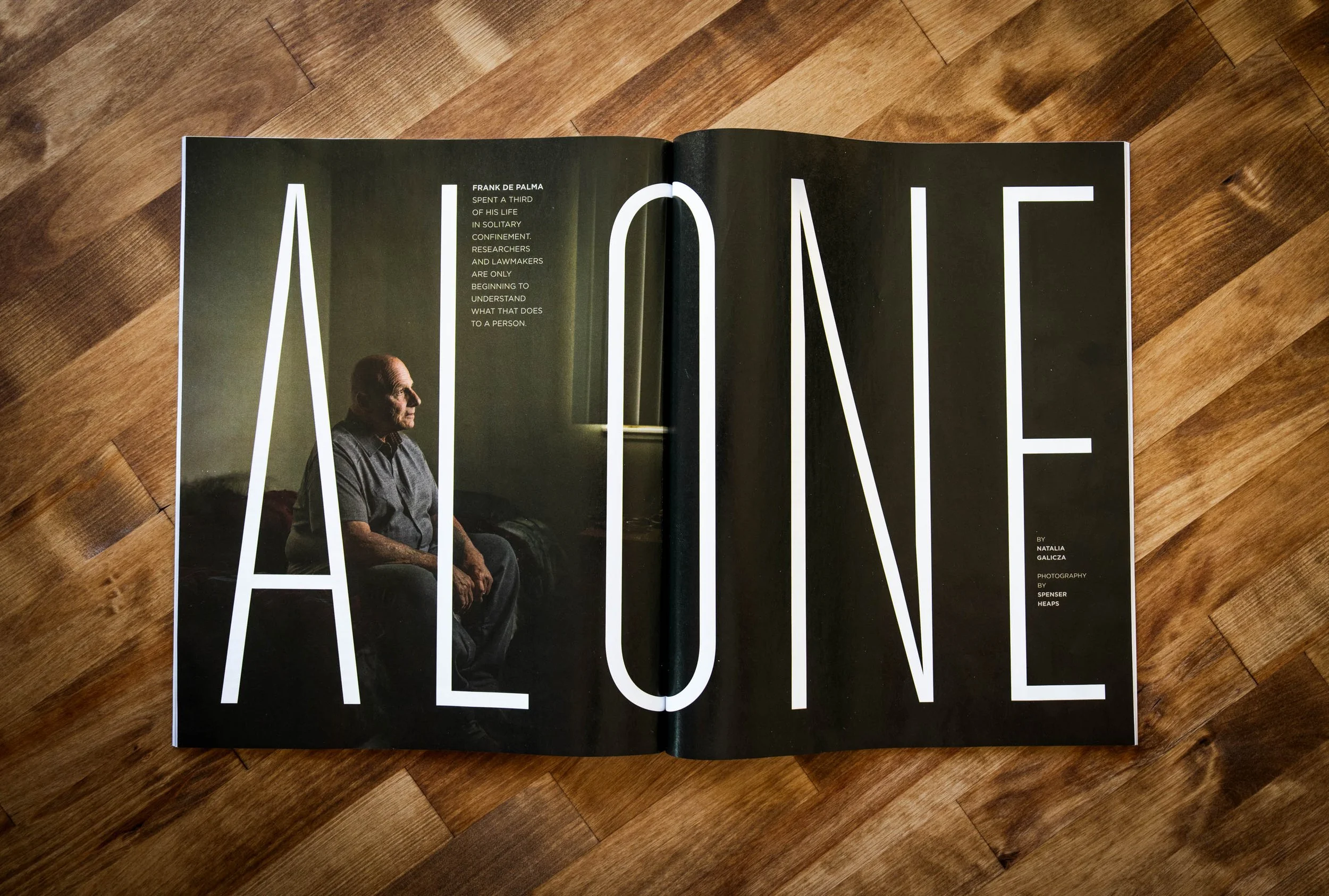 Open magazine featuring a portrait of an elderly man sitting on a chair, with the large word 'ALONE' spanned across both pages in white letters on a dark background.