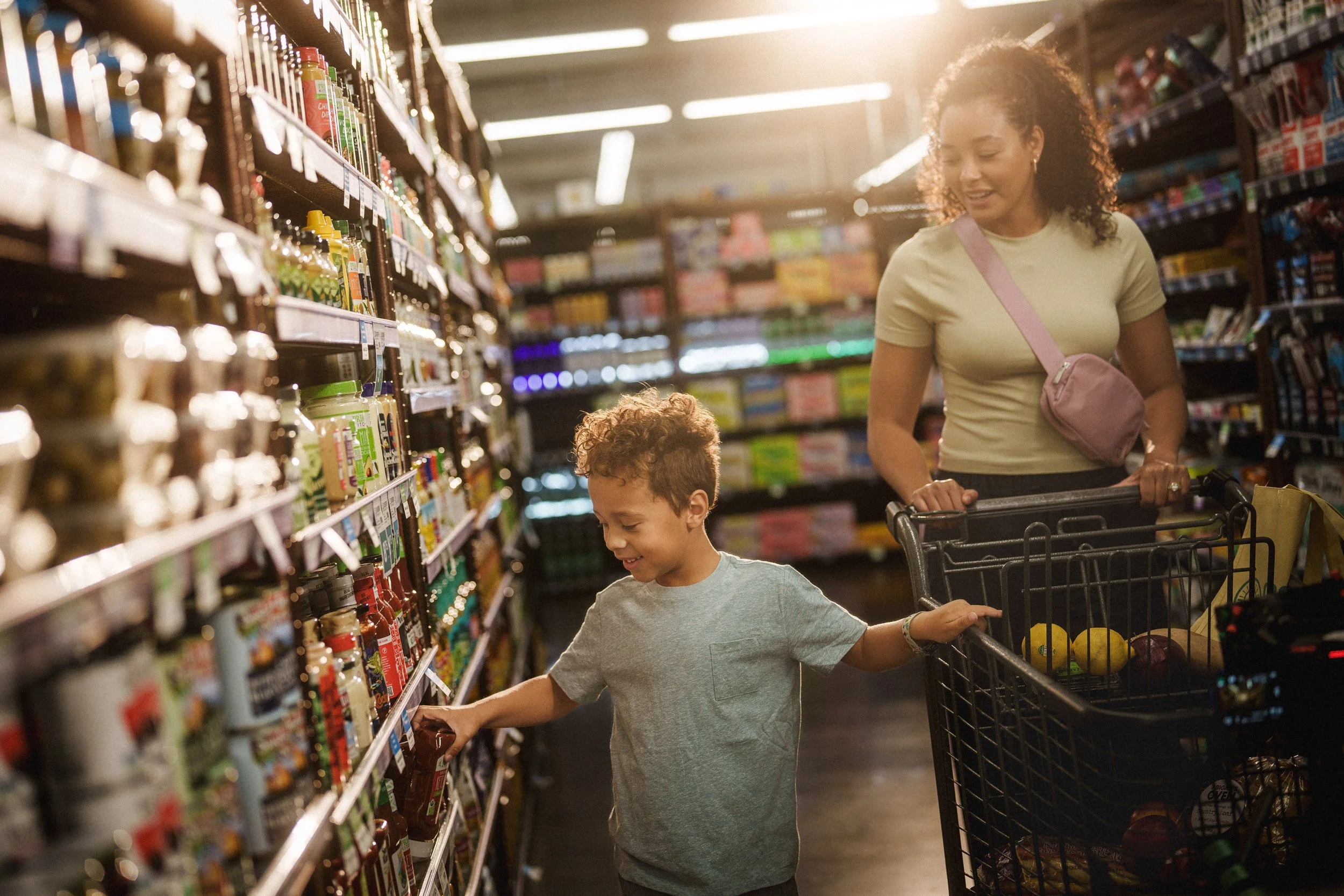 A woman and a young boy shopping for groceries in a supermarket aisle, with the boy reaching for a bottle on the shelf and the woman pushing a shopping cart filled with produce.