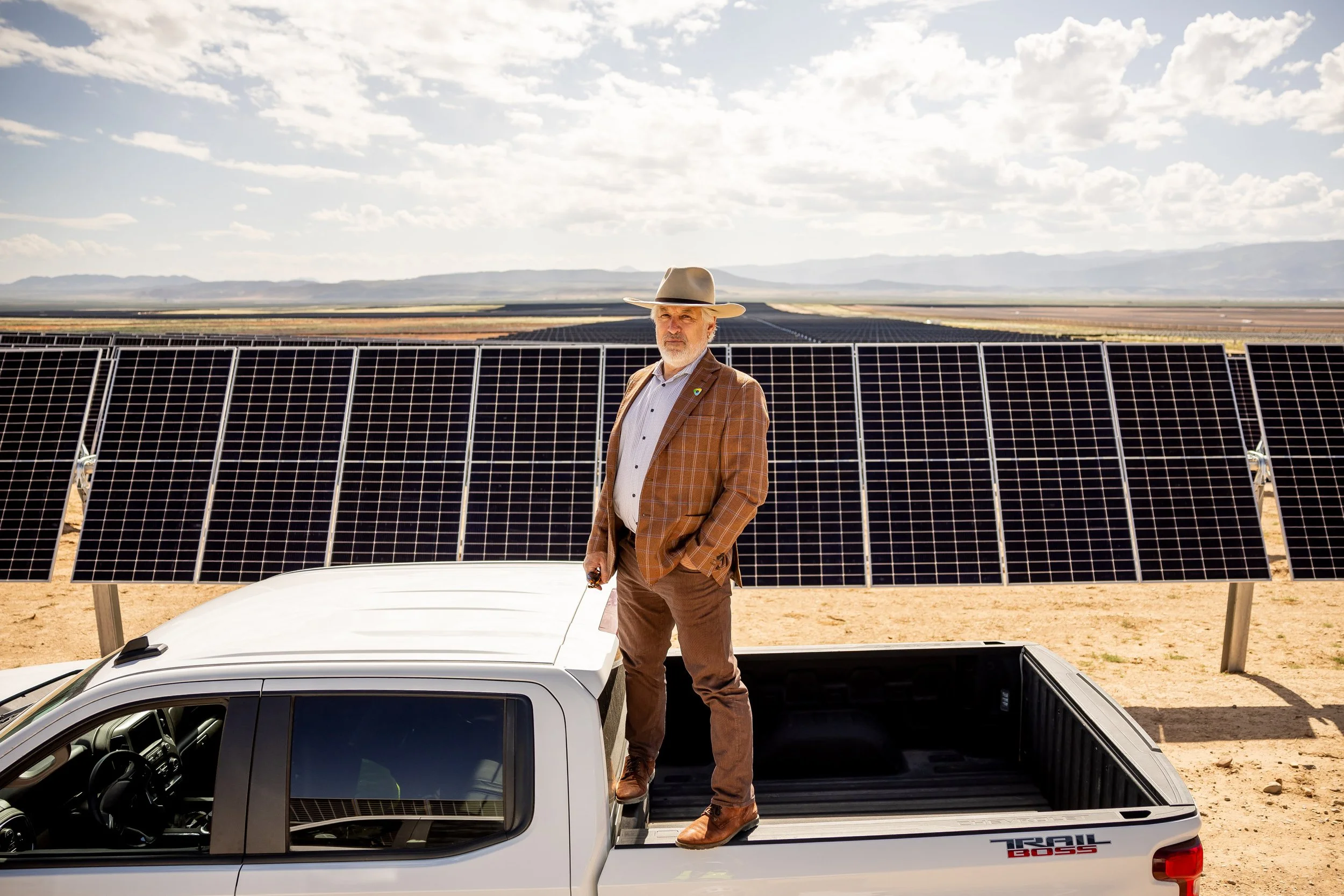 A man in a brown checkered blazer and hat standing in the truck bed of a white pickup truck, with solar panels in the background under a partly cloudy sky.