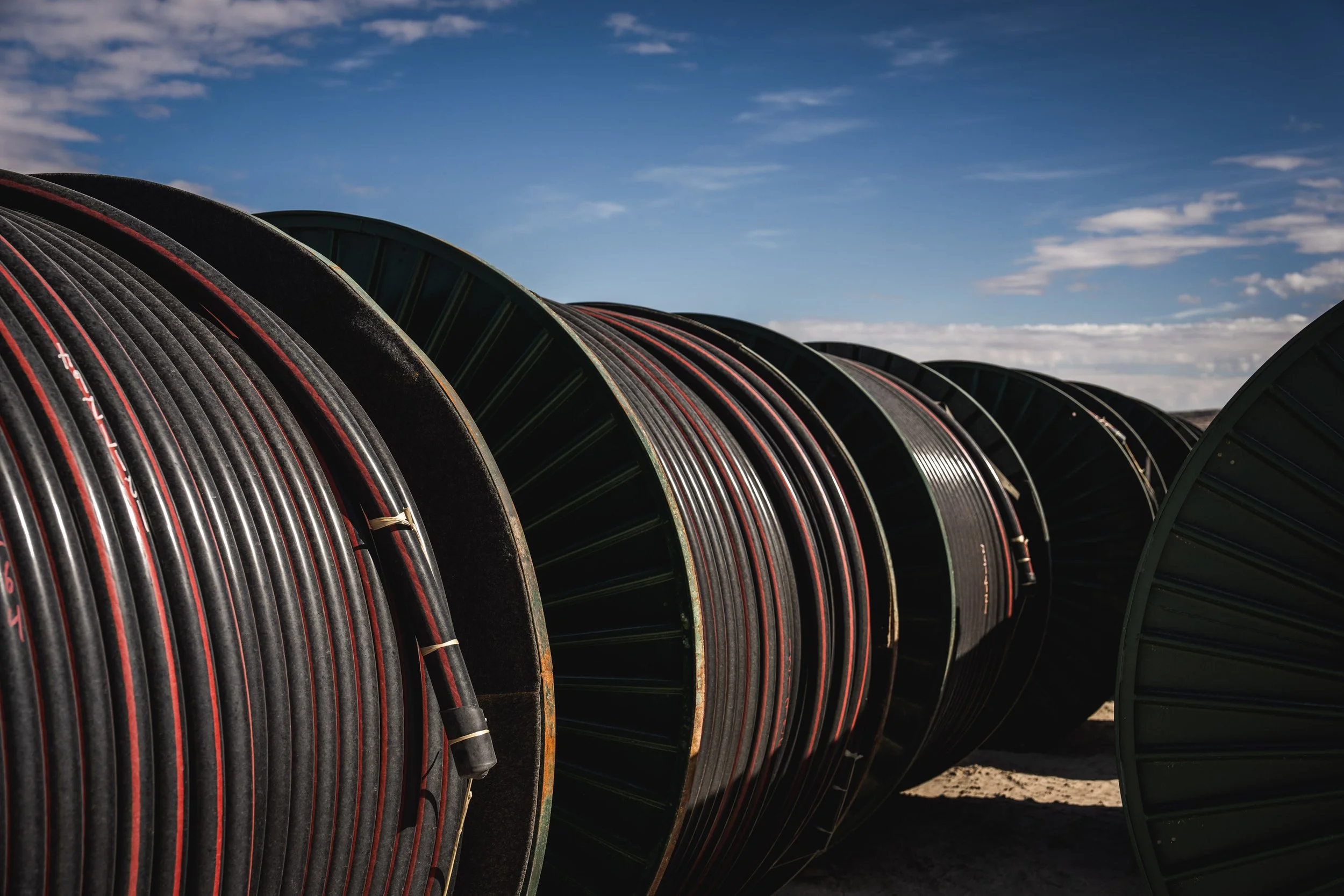 Large spools of black cable stacked outdoors with a blue sky and scattered clouds in the background.