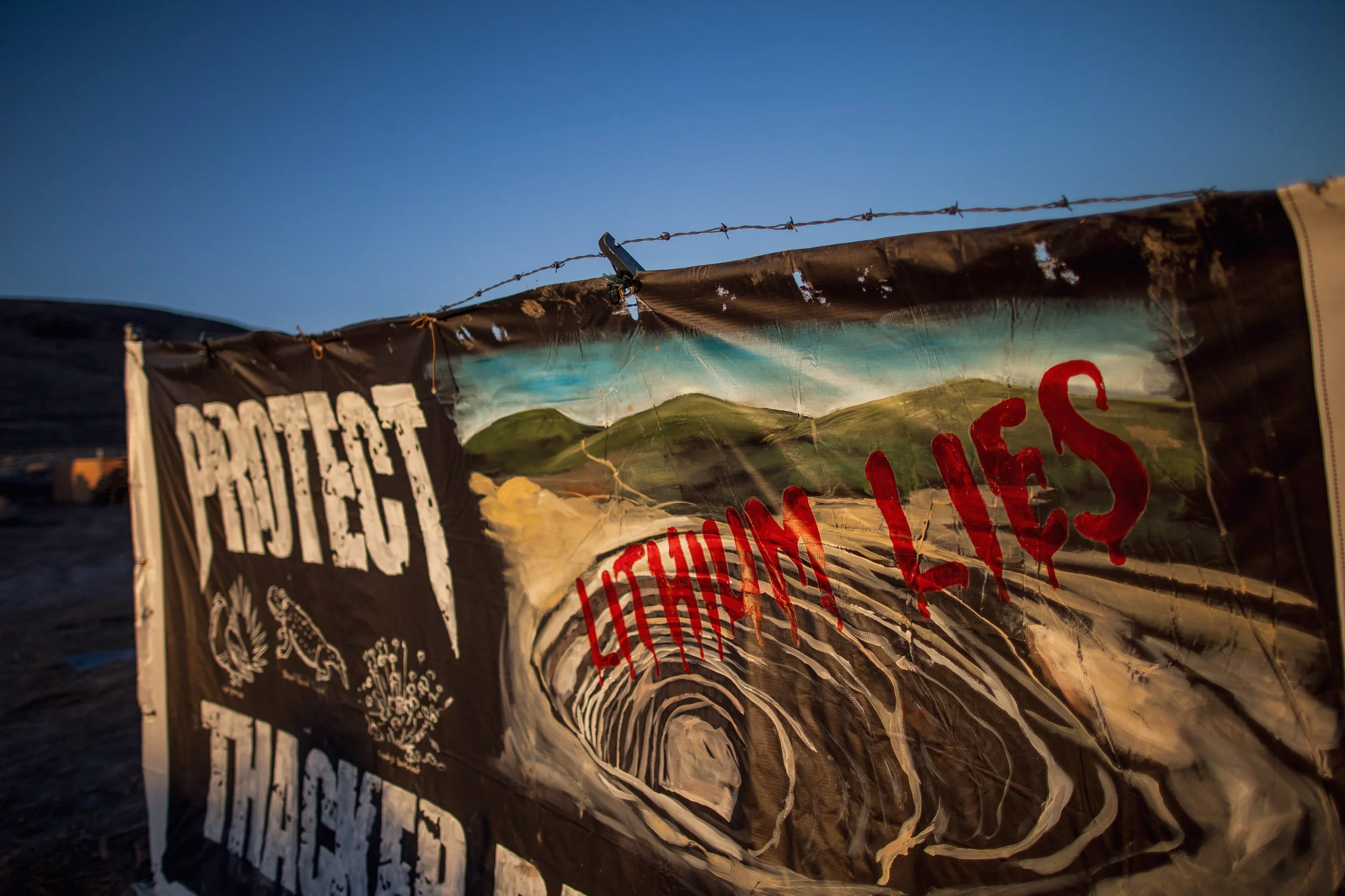 A protest banner with painted landscape, the words "PROTECT THE LAND" in white and red, and messages about land rights, held up by a barbed wire fence during sunset.