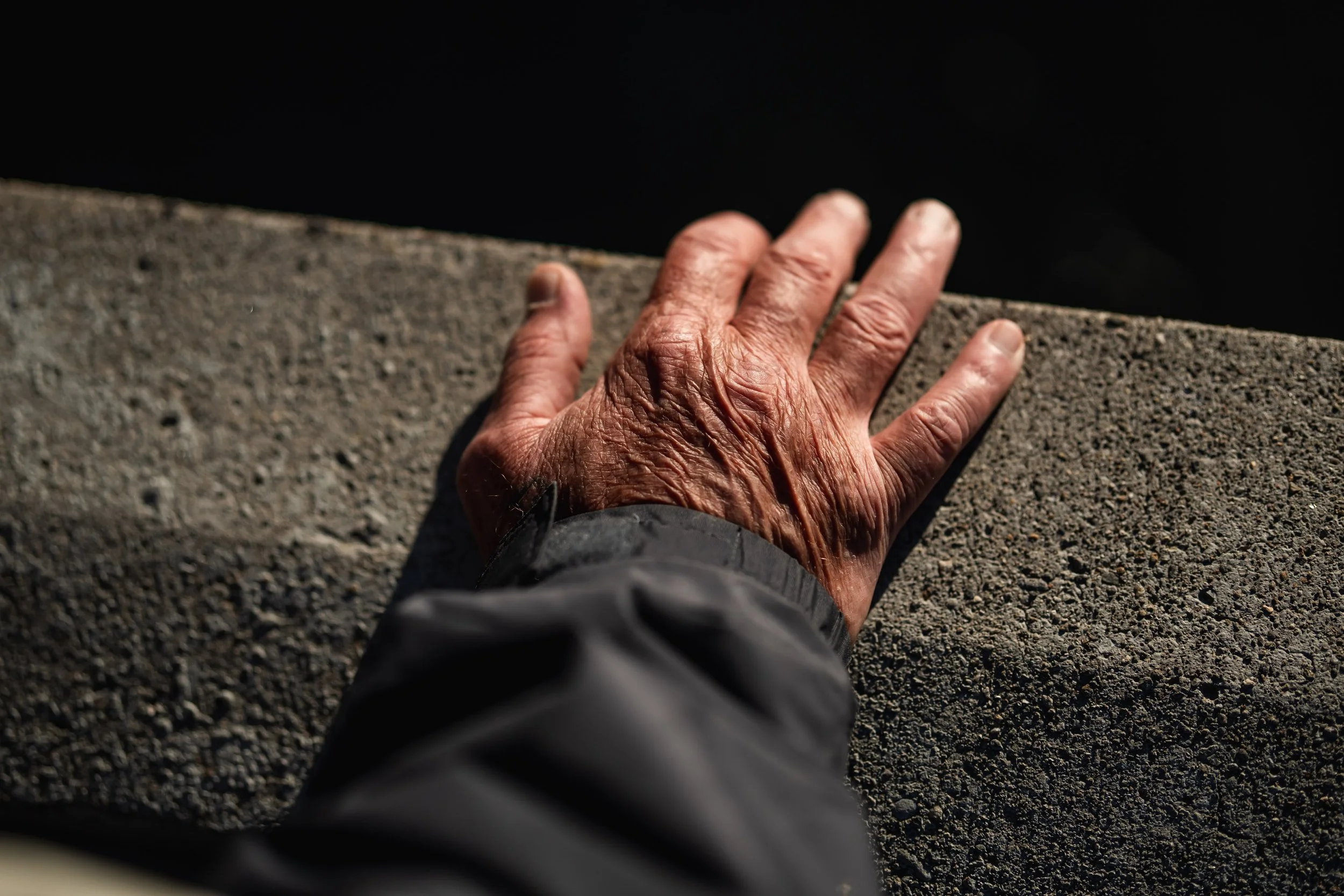 A weathered hand with prominent wrinkles resting on a rough concrete ledge or wall, with a dark blurred background.