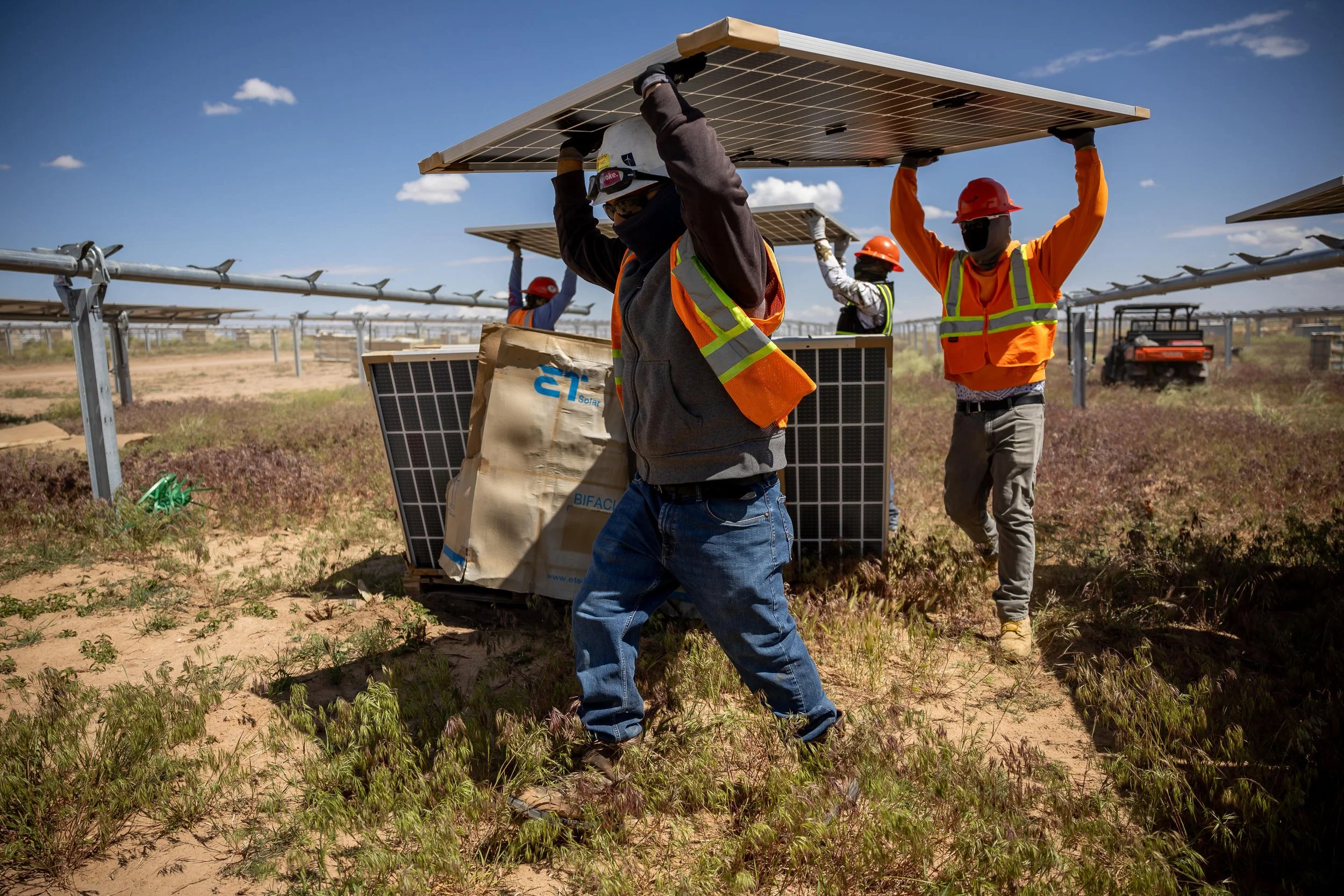 Workers wearing safety vests, masks, helmets, and gloves are installing solar panels in an outdoor solar farm during the daytime.