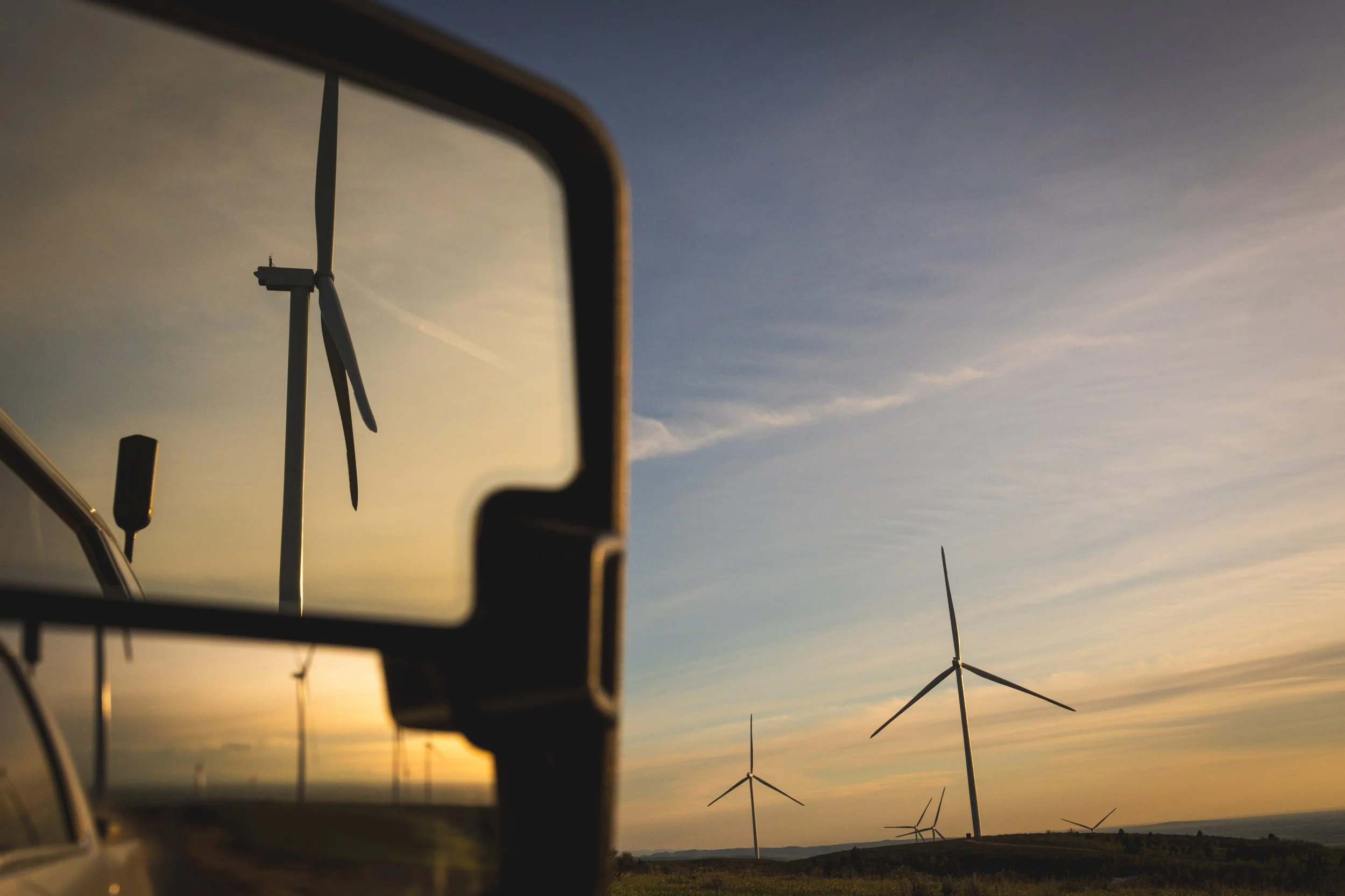 Wind turbines on a hill with a sunset sky, seen through a vehicle's side mirror.