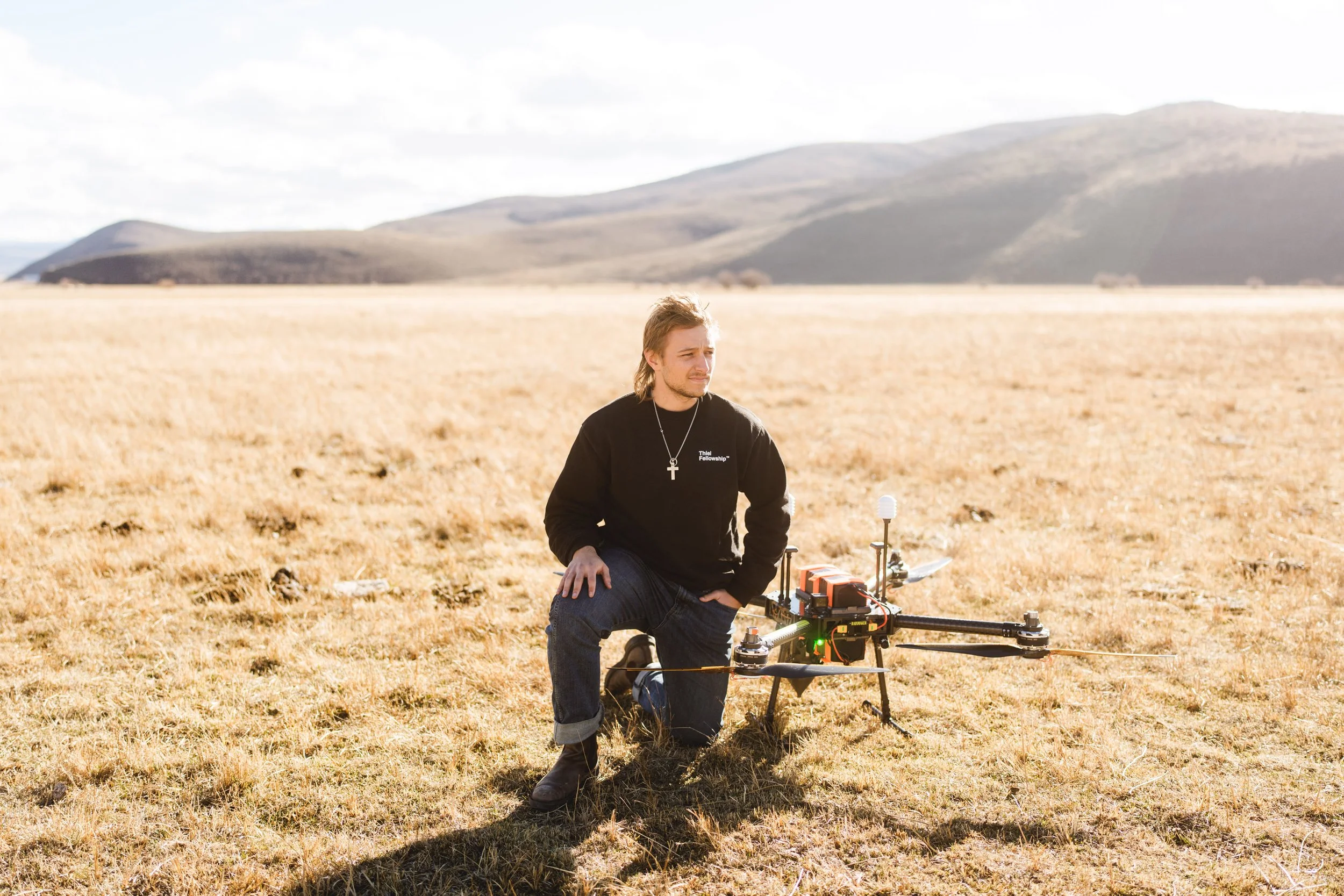 A man kneeling on the grass in an open field with mountains in the background, next to a large drone with multiple propellers and equipment mounted on it.