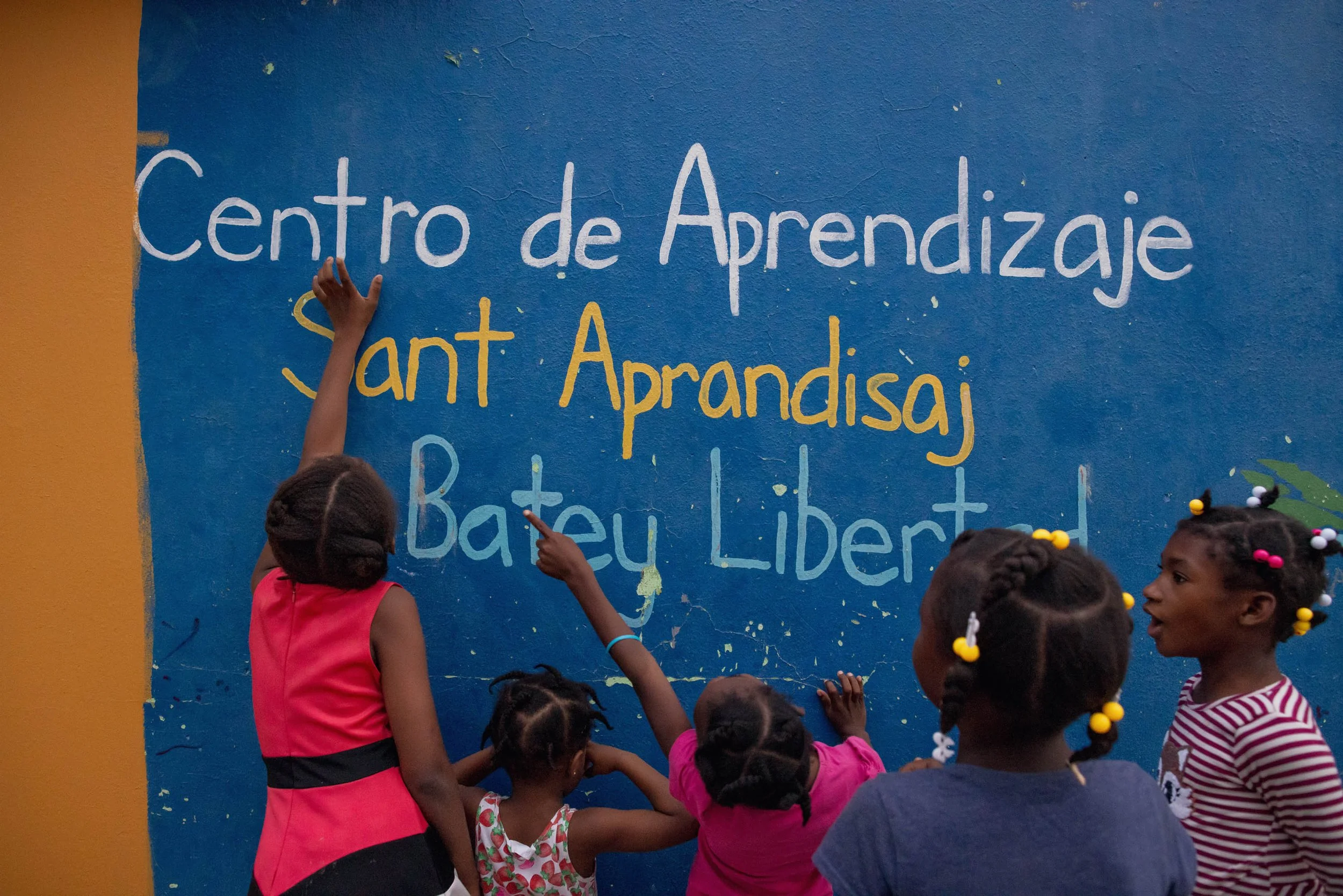 Children gathered in front of a blue wall with Spanish and Haitian Creole writing, some pointing and one reaching to touch the wall.