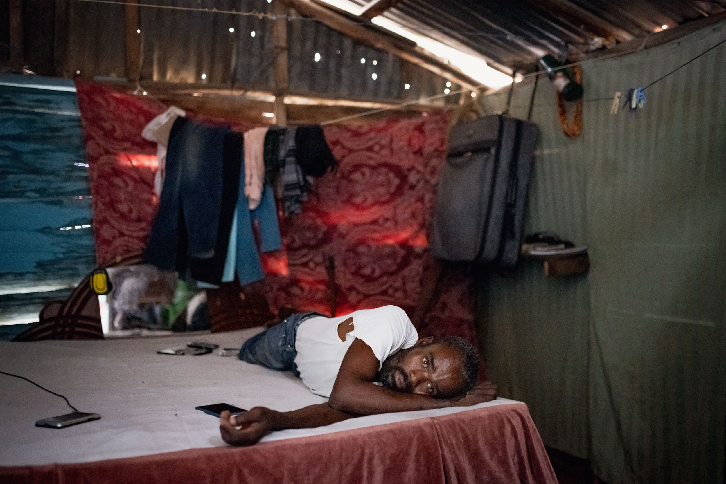 A man with a beard and mustache lying on a bed in a modest room, propped up on his side, looking at the camera. The room has hanging clothes, a suitcase, and a small shelf with items, with a tin roof and wooden walls.