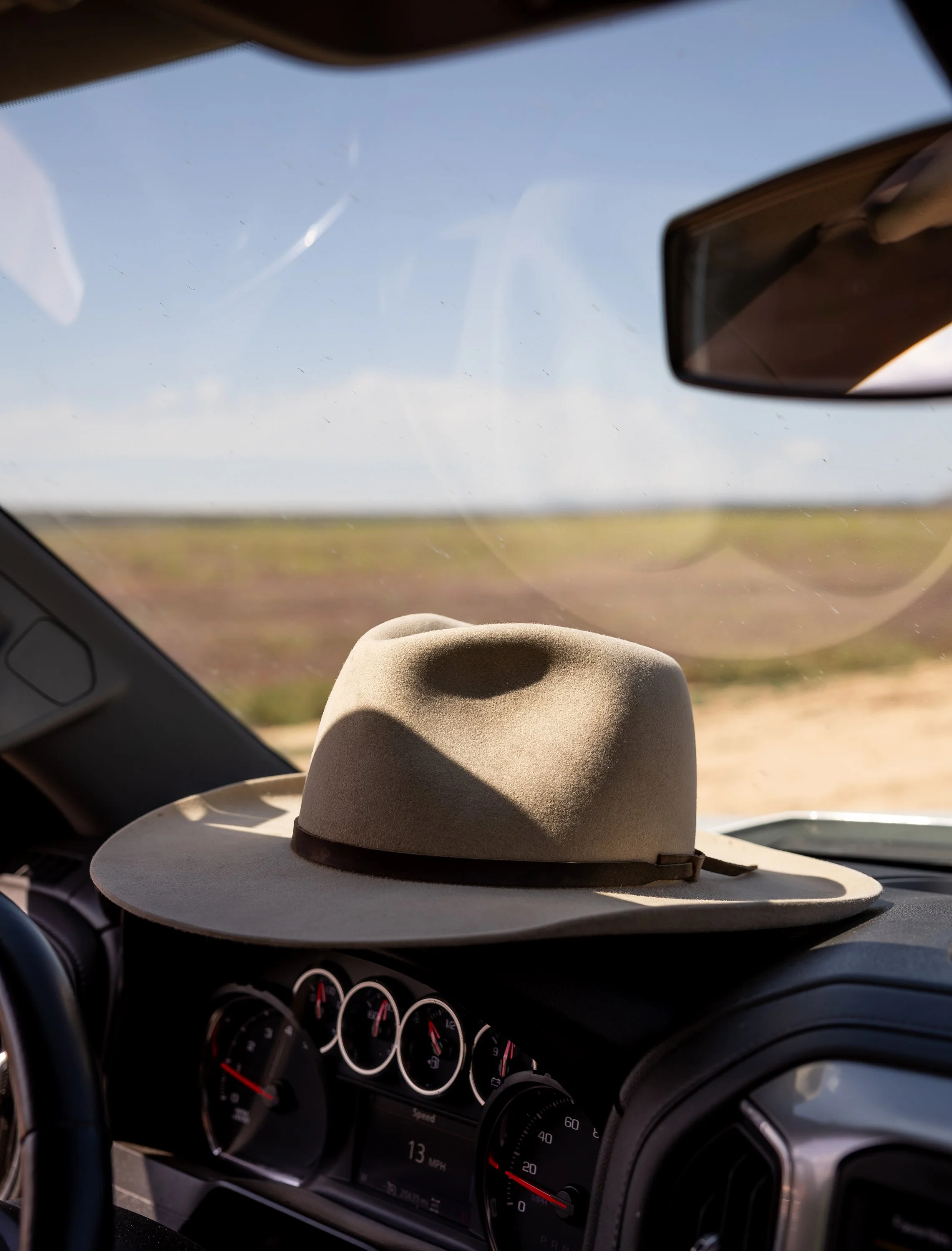 A beige cowboy hat resting on the dashboard of a car with a desert landscape visible through the windshield.