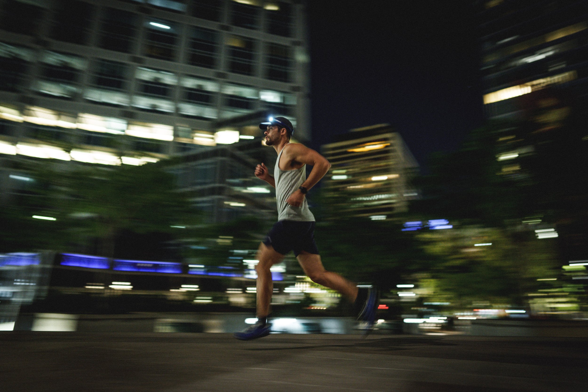 A man running at night in an urban area with tall buildings and illuminated windows behind him, motion blur showing speed.
