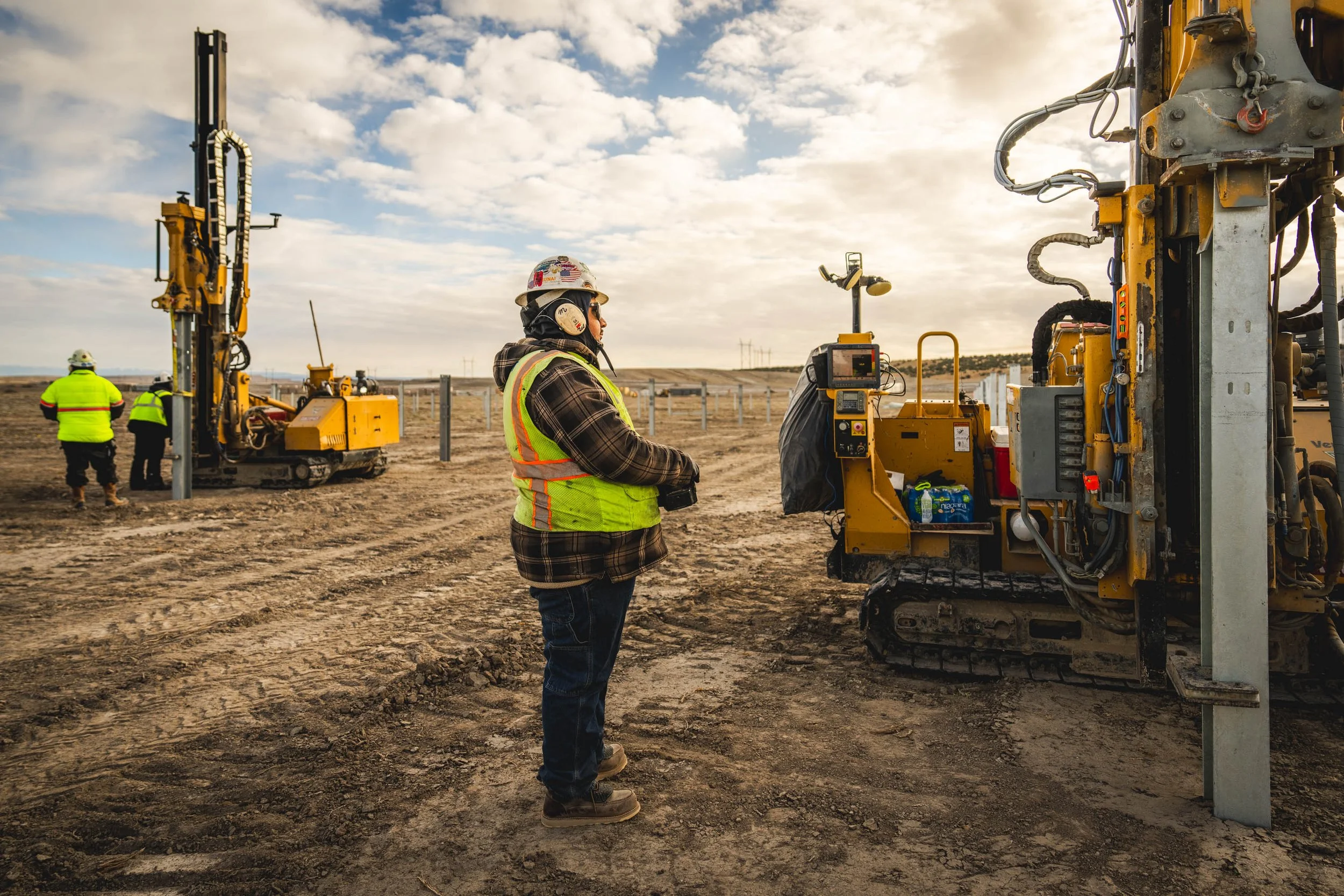 Construction workers at a solar energy project site operating machinery and inspecting equipment under a partly cloudy sky.