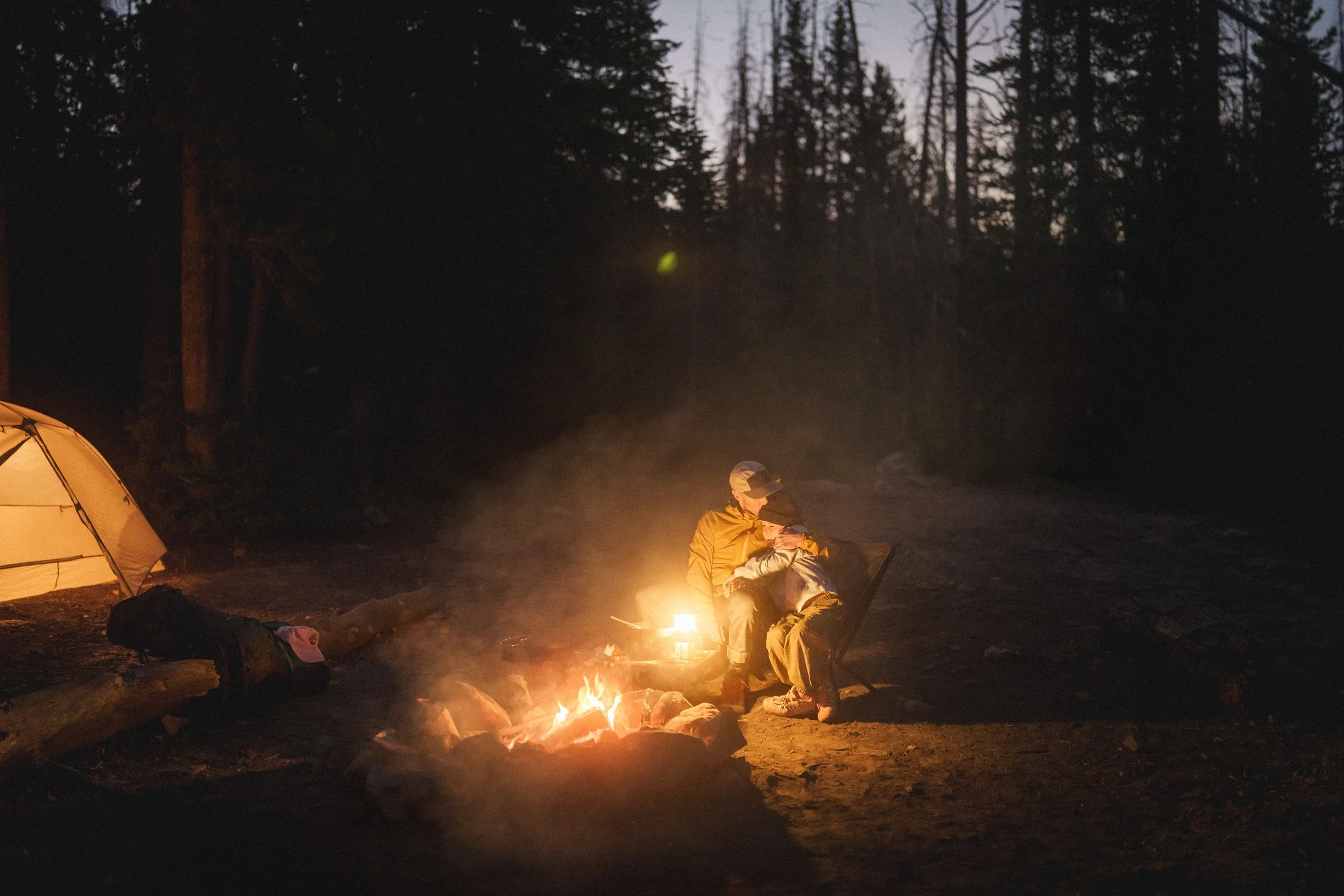 People sitting around a campfire at night in a wooded area, with a tent nearby.
