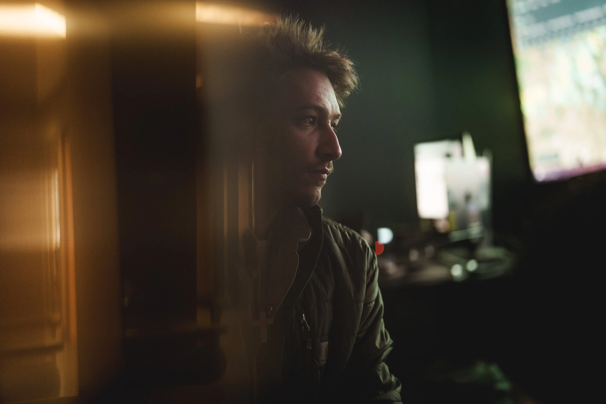 A young man with blonde hair sitting in a dimly lit room, looking at a computer screen with multiple monitors.