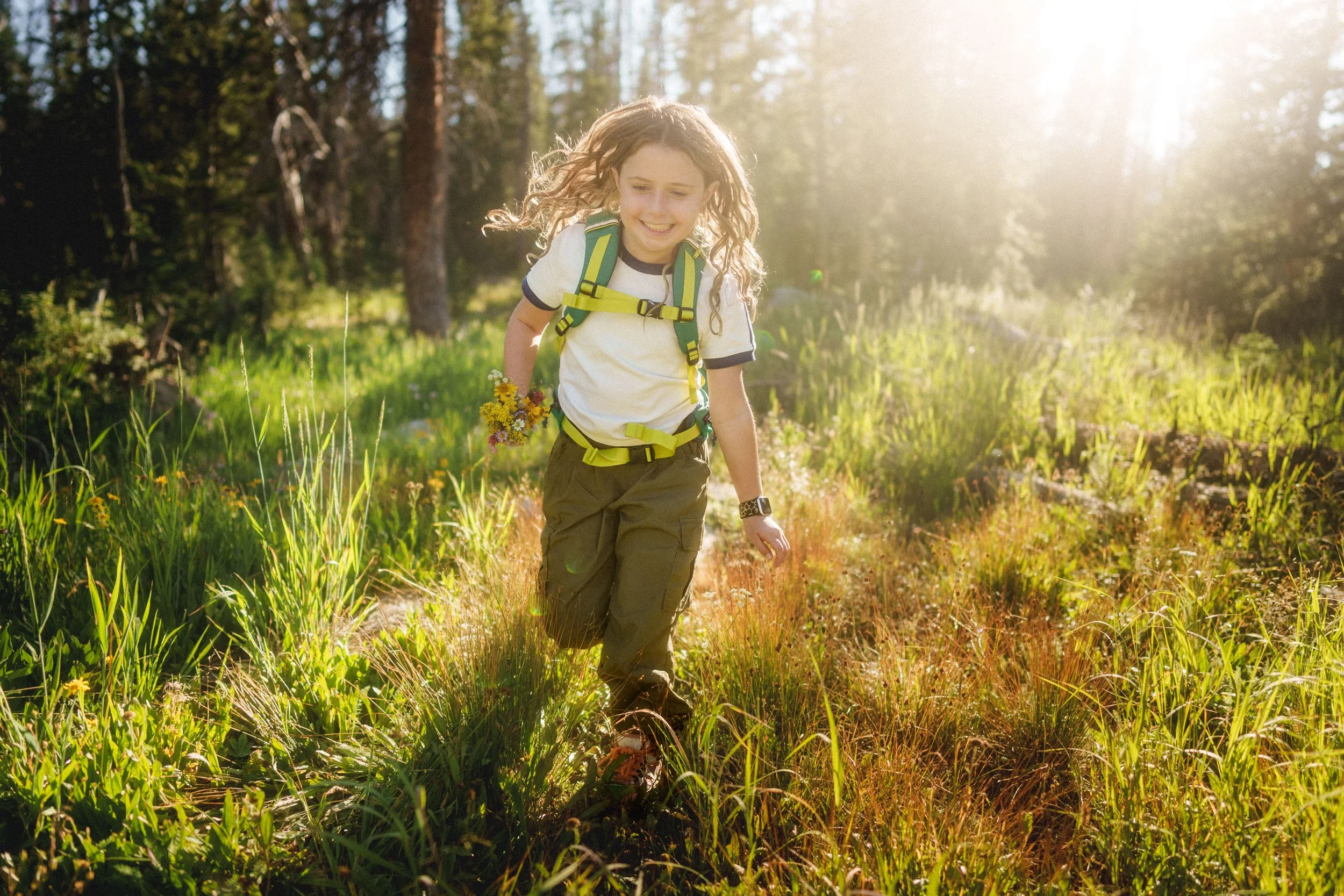 Young girl hiking through a sunlit forest trail, carrying a small bouquet of wildflowers, wearing a backpack and outdoor clothing.