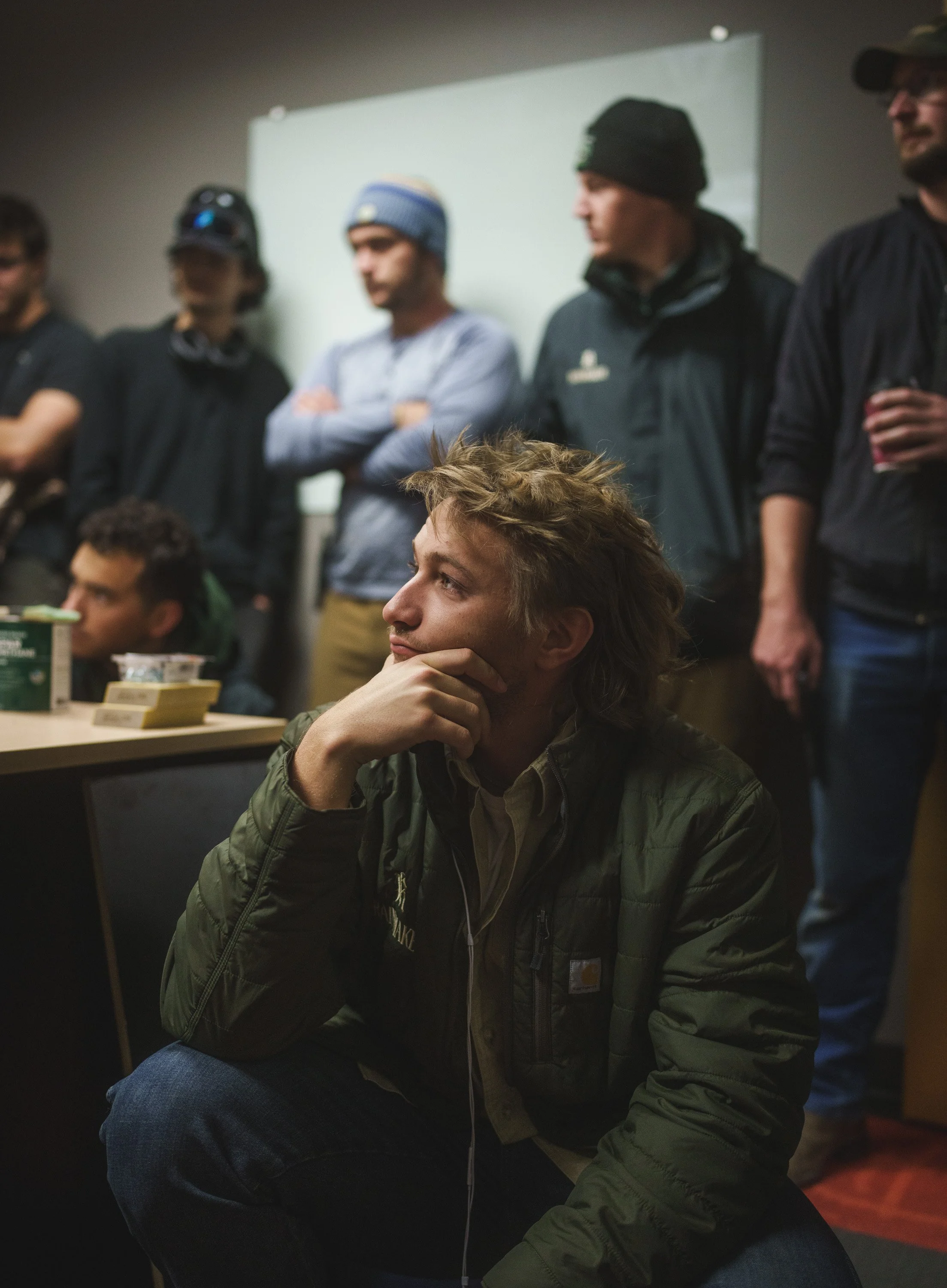A group of men gathered in a room, with one man sitting and deep in thought with his hand on his chin, while others stand behind him, some with arms crossed and holding a drink.