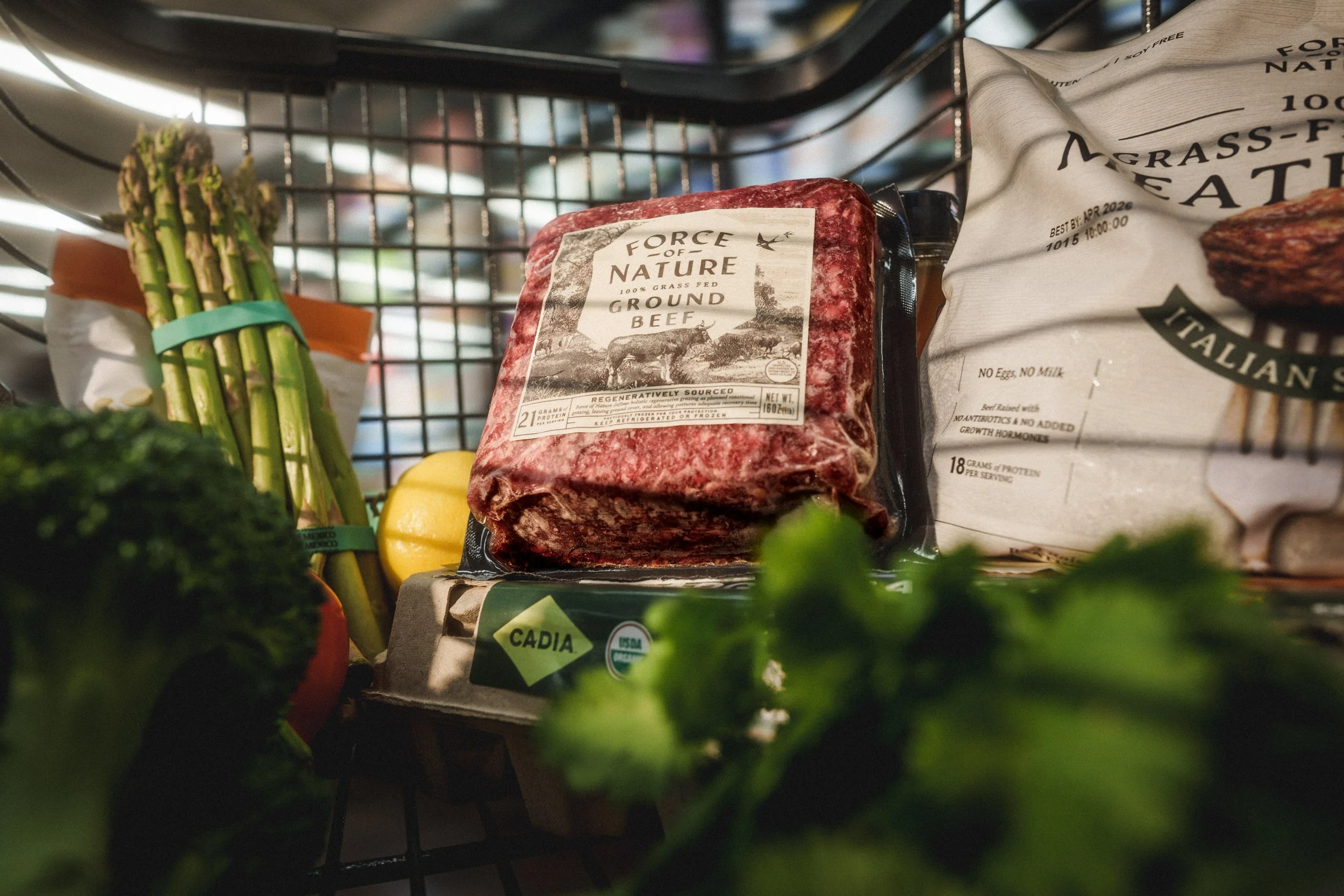 A grocery cart containing a package of ground beef, a bag of Italian bread, and fresh vegetables including asparagus, broccoli, and a lemon.