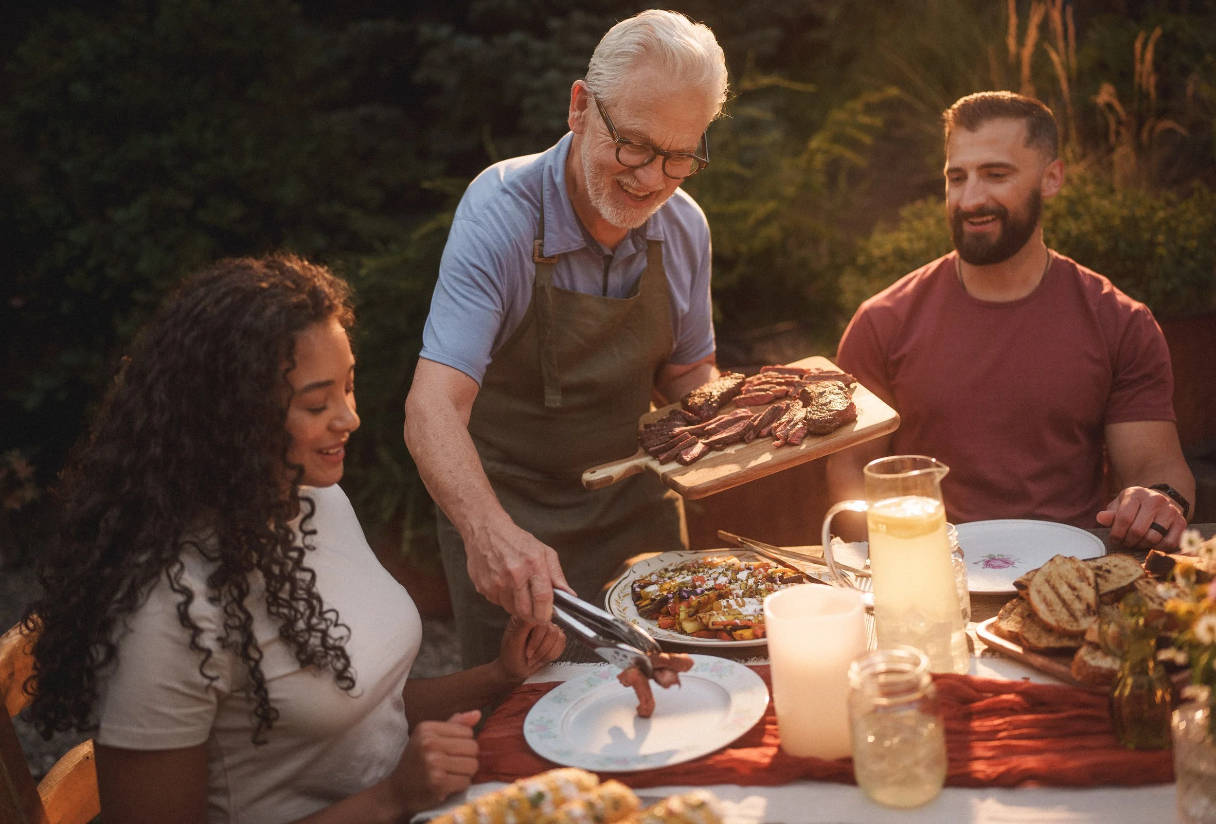 People enjoying an outdoor dinner party with a person serving grilled meats, a woman eating, and a man sitting at the table, with food and drinks on the table during sunset.