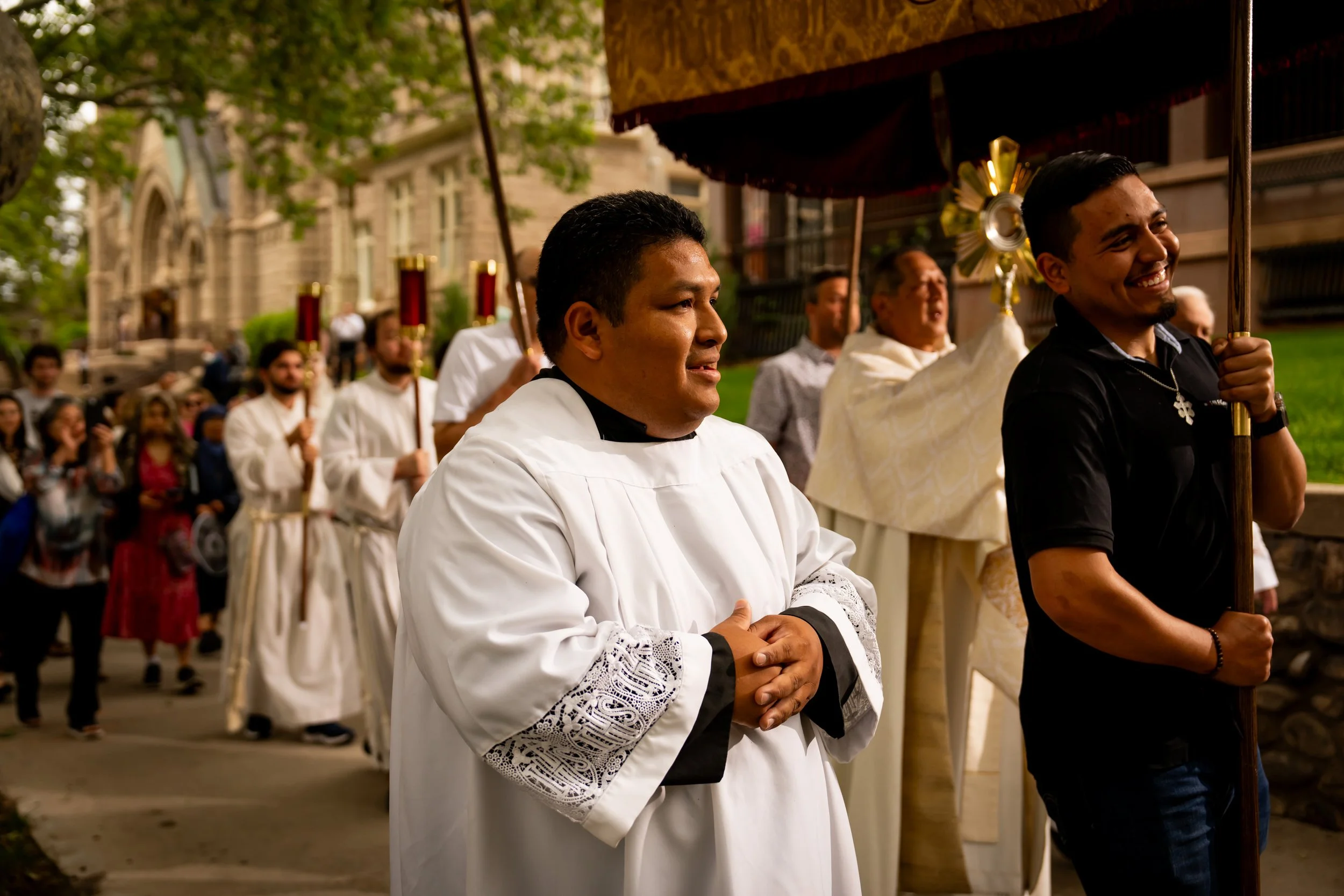 A church procession with people dressed in religious robes, walking outdoors on a church street, with some participants carrying candles and others holding a canopy over a priest or religious leader.