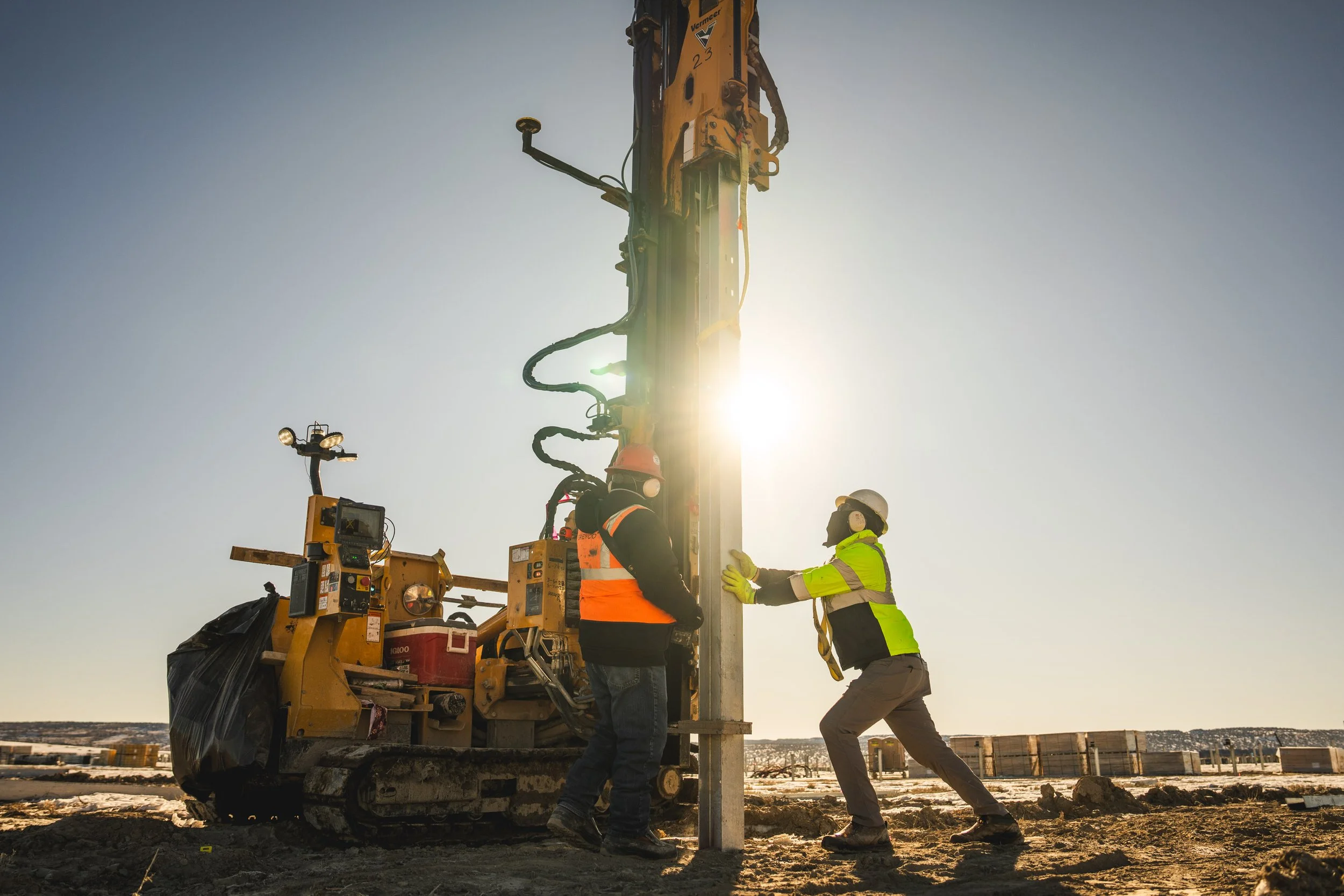 Two construction workers wearing safety gear and high-visibility vests installing or maintaining a utility pole using heavy machinery on a construction site during daylight.
