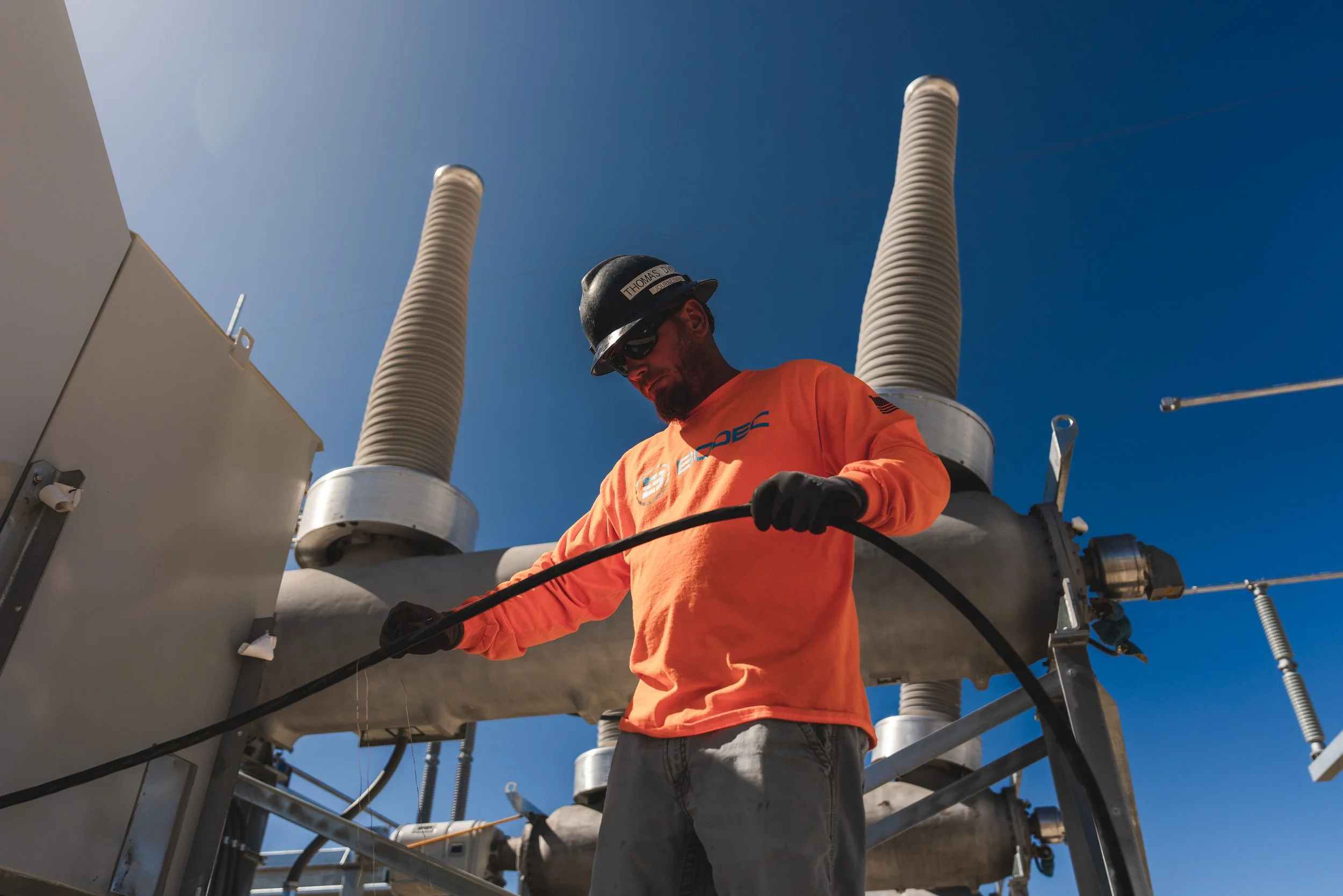 A worker in an orange shirt and black helmet is handling a cable in front of large electrical insulators and equipment outdoors under a clear blue sky.