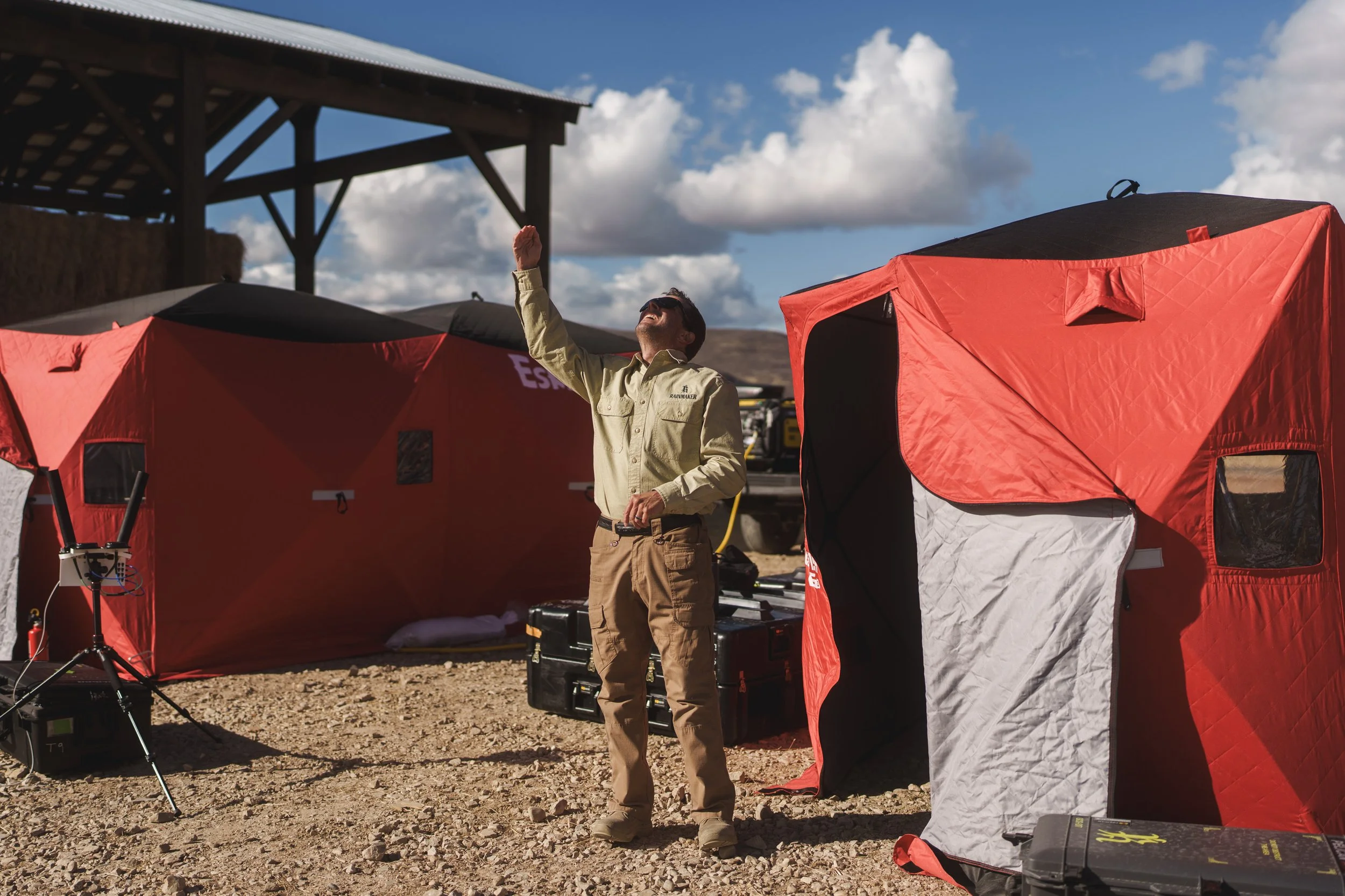 A man standing outside among red and white temporary structures, raising his arm and looking up at the sky with a smile.